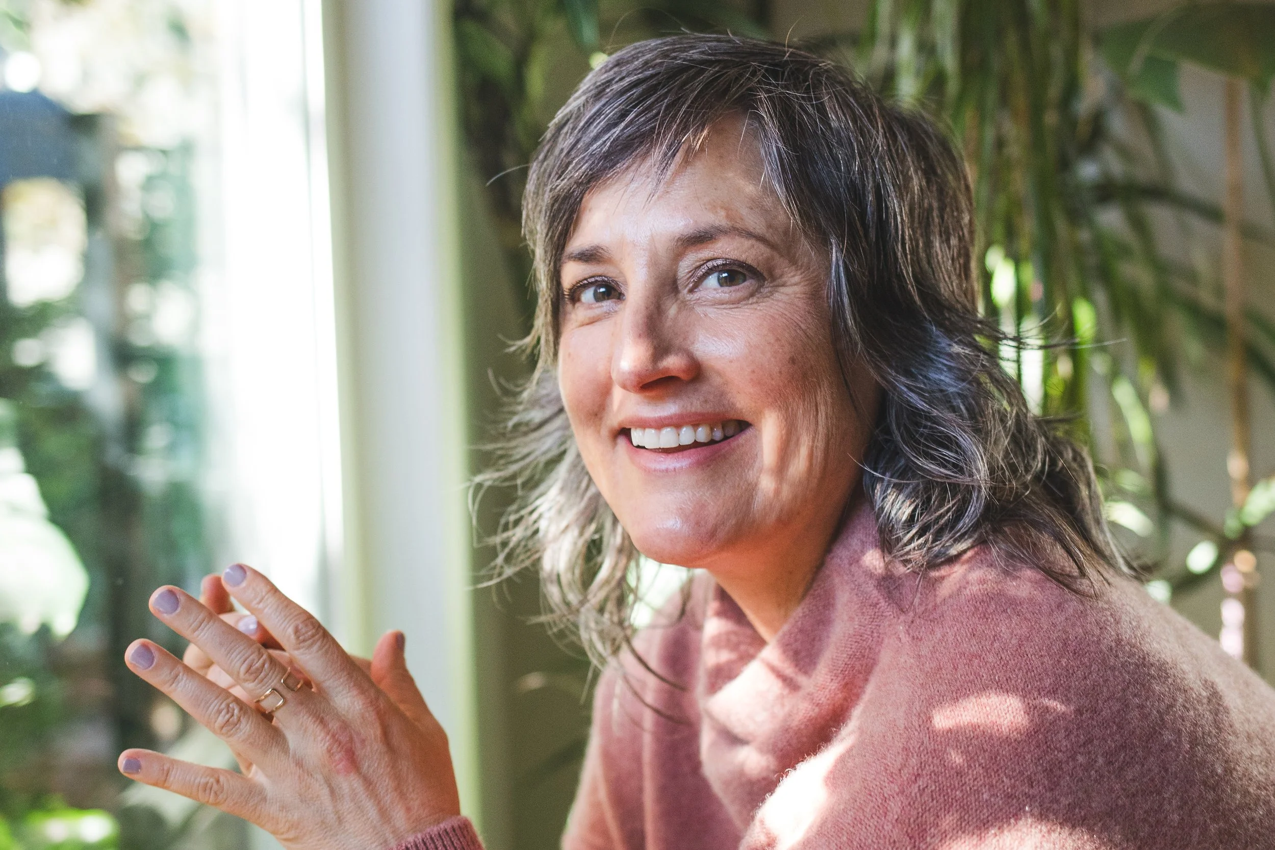 Pema smiling while looking slightly to the side, sitting indoors with a background of green plants and sunlight.