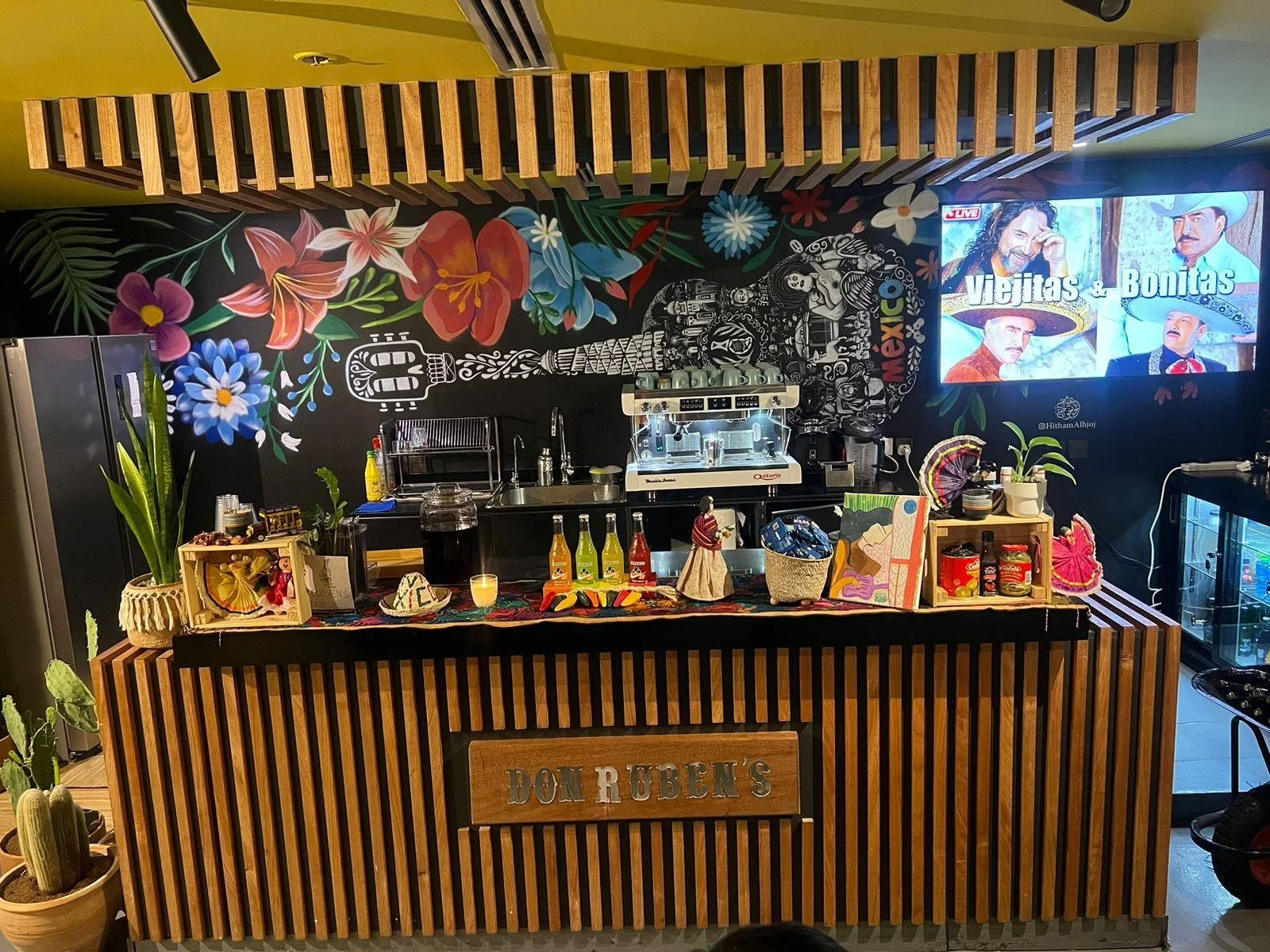 Barra con decoraciones mexicanas, mural de flores y guitarra, pantallas con imágenes de mariachi y la leyenda "Viejitas y Bonitas".
