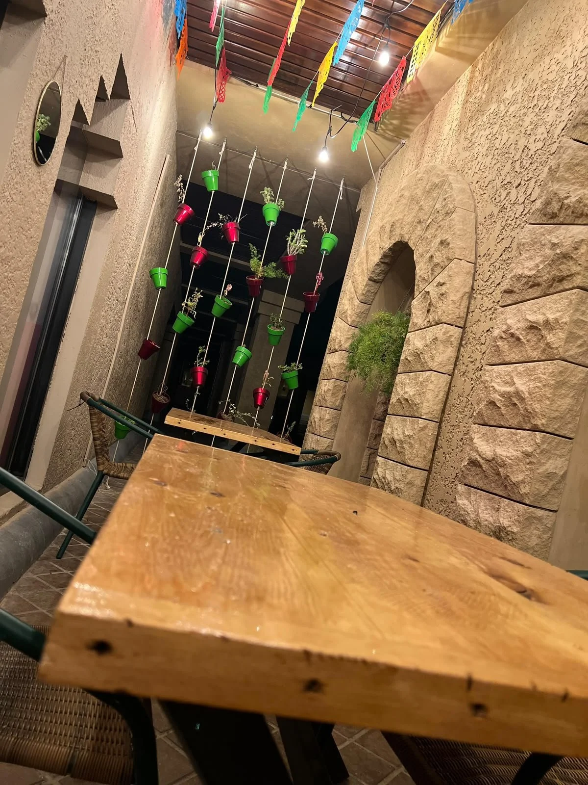 Muebles de madera en un espacio decorativo con paredes de piedra, colgantes con macetas de diferentes colores con plantas y banderines de papel de colores en el techo.