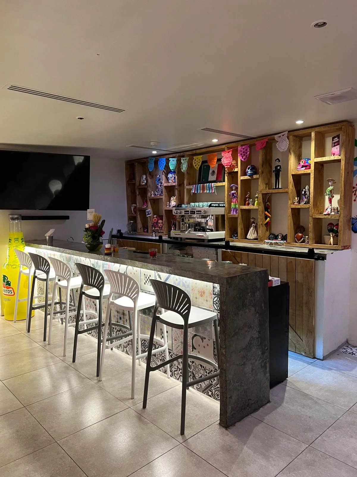 Barra de un restaurante con cinco sillas blancas y negras, decorada con colores vivos y objetos culturales mexicanos, fondo con estanterías de madera llenas de objetos decorativos tradicionales y una máquina de café.