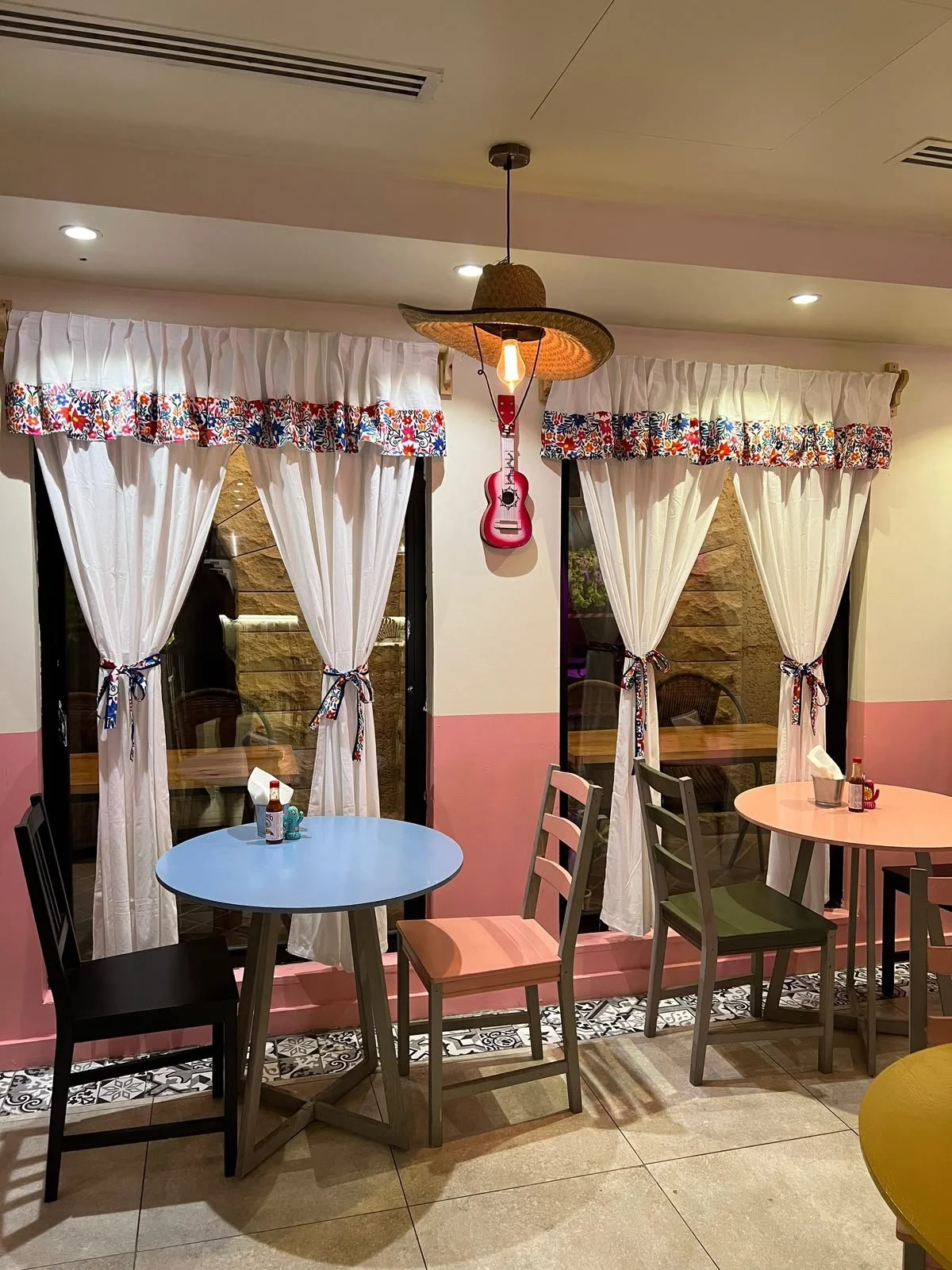 Interior de un restaurante con cortinas blancas con bordes de colores, mesas redondas de colores pastel, sillas de diferentes colores, un tocadiscos decorativo en la pared y una lámpara de sombrero en el techo.