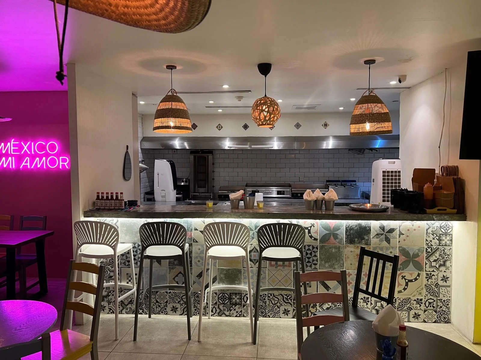 Interior de un restaurante con barra y sillas blancas. Decoración con azulejos coloridos en la barra y luces colgantes de cestería. En el fondo, área de cocina y cartel de neón en la pared izquierda que dice 'México mi amor'.
