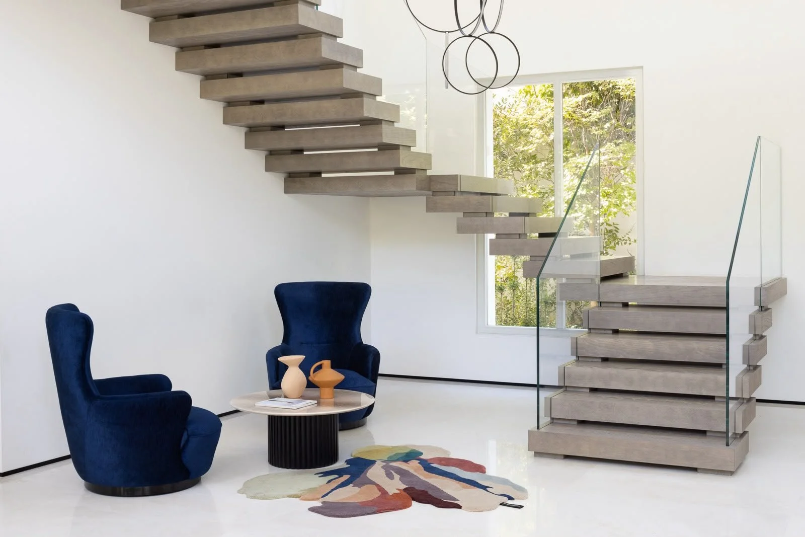 Modern minimalist interior with floating concrete stairs, glass railing, two navy blue velvet chairs, a round coffee table with decorative vases, a colorful abstract rug, and a large window with green trees outside.