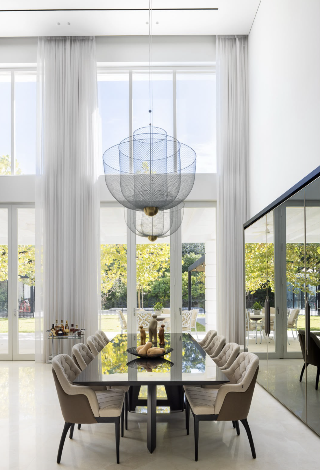 Modern dining room with a black table, cream upholstered chairs, and decorative figurines. Large windows with white curtains let in natural light, overlooking a garden with trees. A contemporary chandelier hangs from the high ceiling.