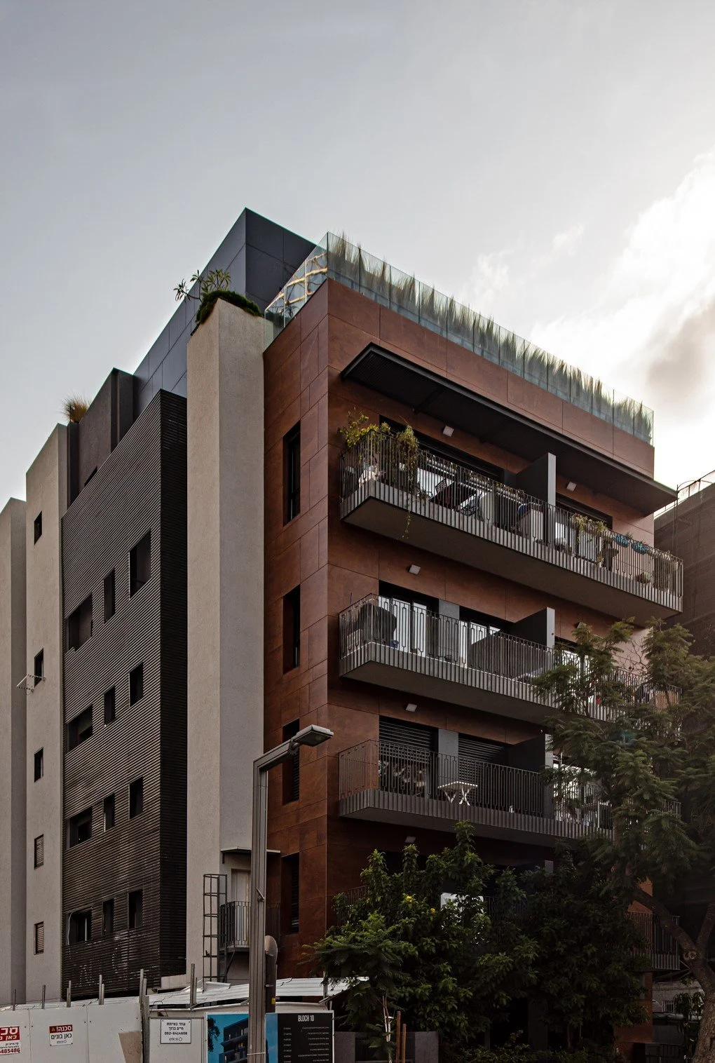 Modern multi-story residential building with balconies, trees, and streetlamp in foreground.