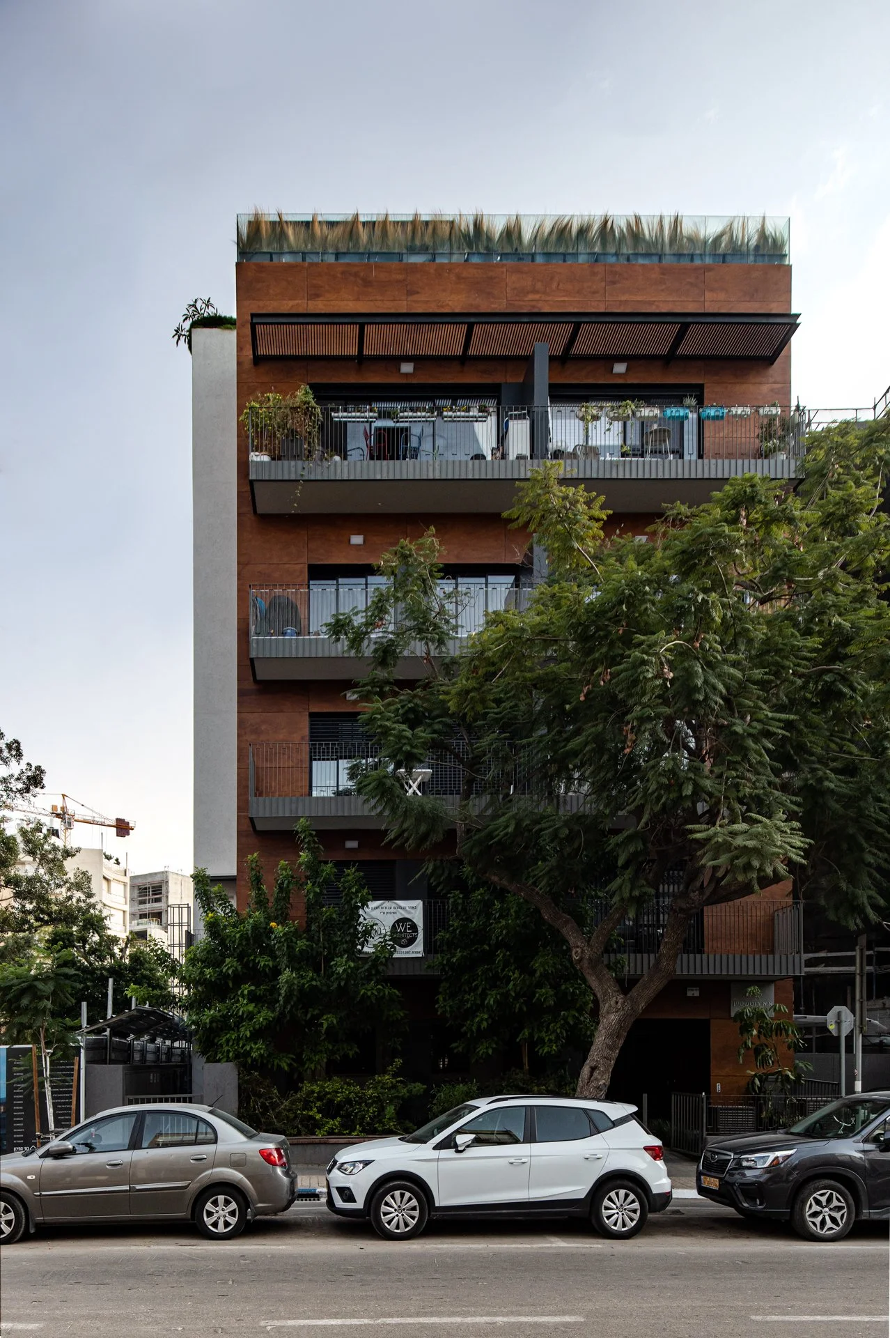 Multi-story modern residential building with balconies, trees in front, parked cars on the street.