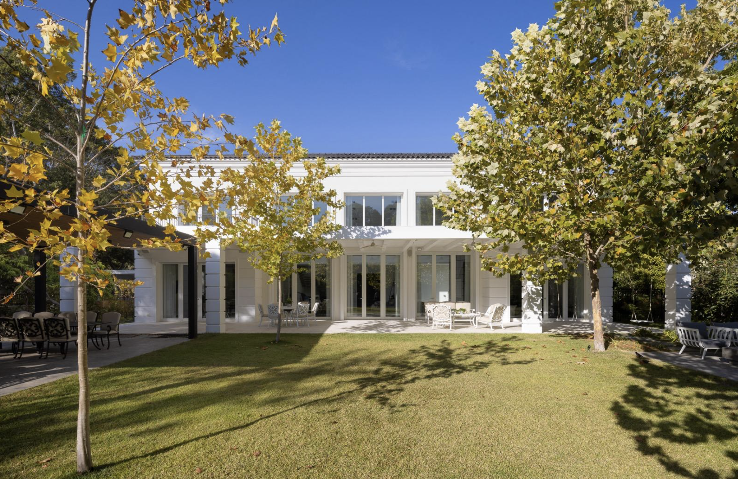 Backyard with green lawn, several trees with yellow-green leaves, patio furniture, and a white two-story house with large windows and patio doors under a clear blue sky.