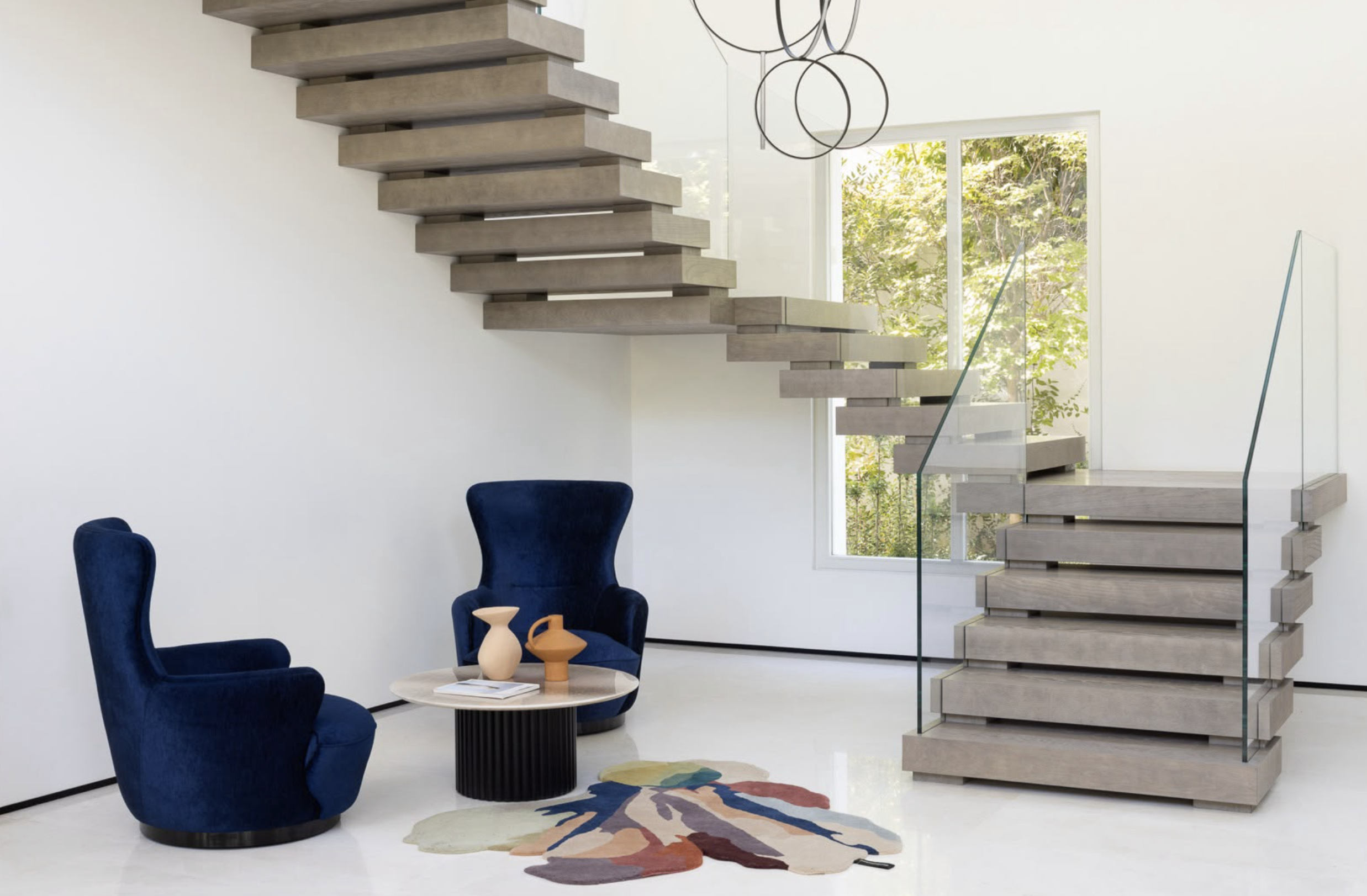 Modern interior with a floating staircase, two dark blue armchairs with high backs, a round coffee table with three vases, colorful pattern rug, large window with trees outside, and black circular chandelier lights.