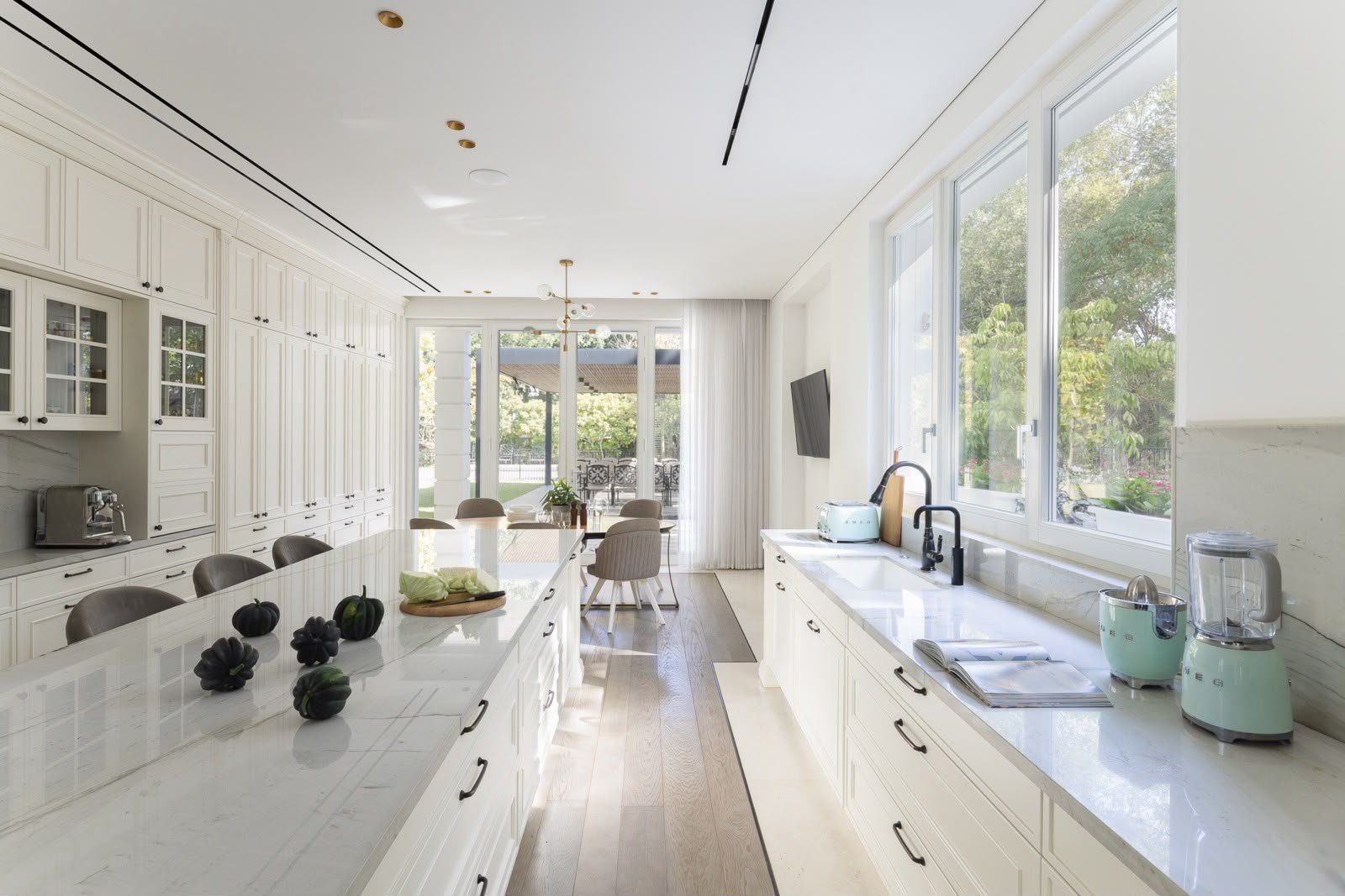Bright modern kitchen with white cabinets, marble countertops, and large windows overlooking a garden.