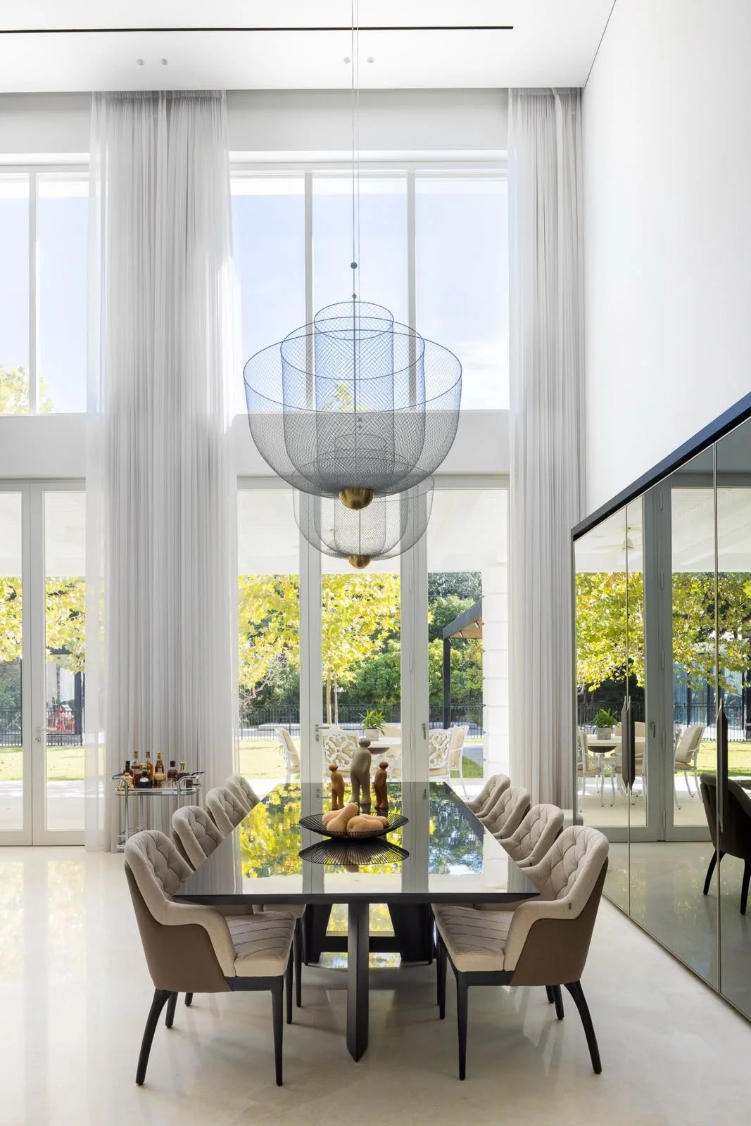 Modern dining room with floor-to-ceiling windows, white curtains, a black dining table surrounded by beige upholstered chairs, and a large, layered wire pendant chandelier; outside view of green trees and patio area.