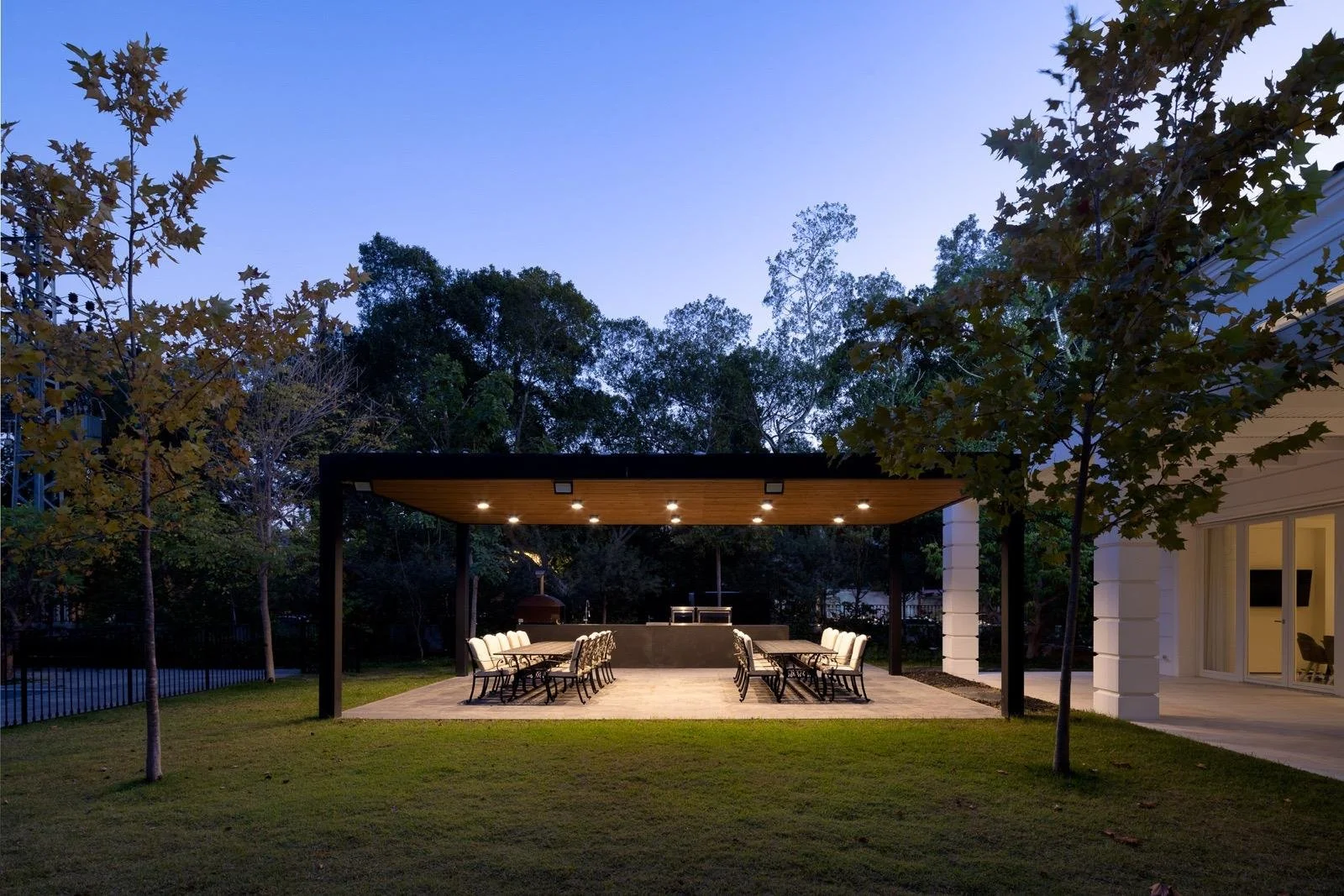 Outdoor patio at twilight with tables and chairs under a wooden canopy, surrounded by trees and a house with sliding glass doors.