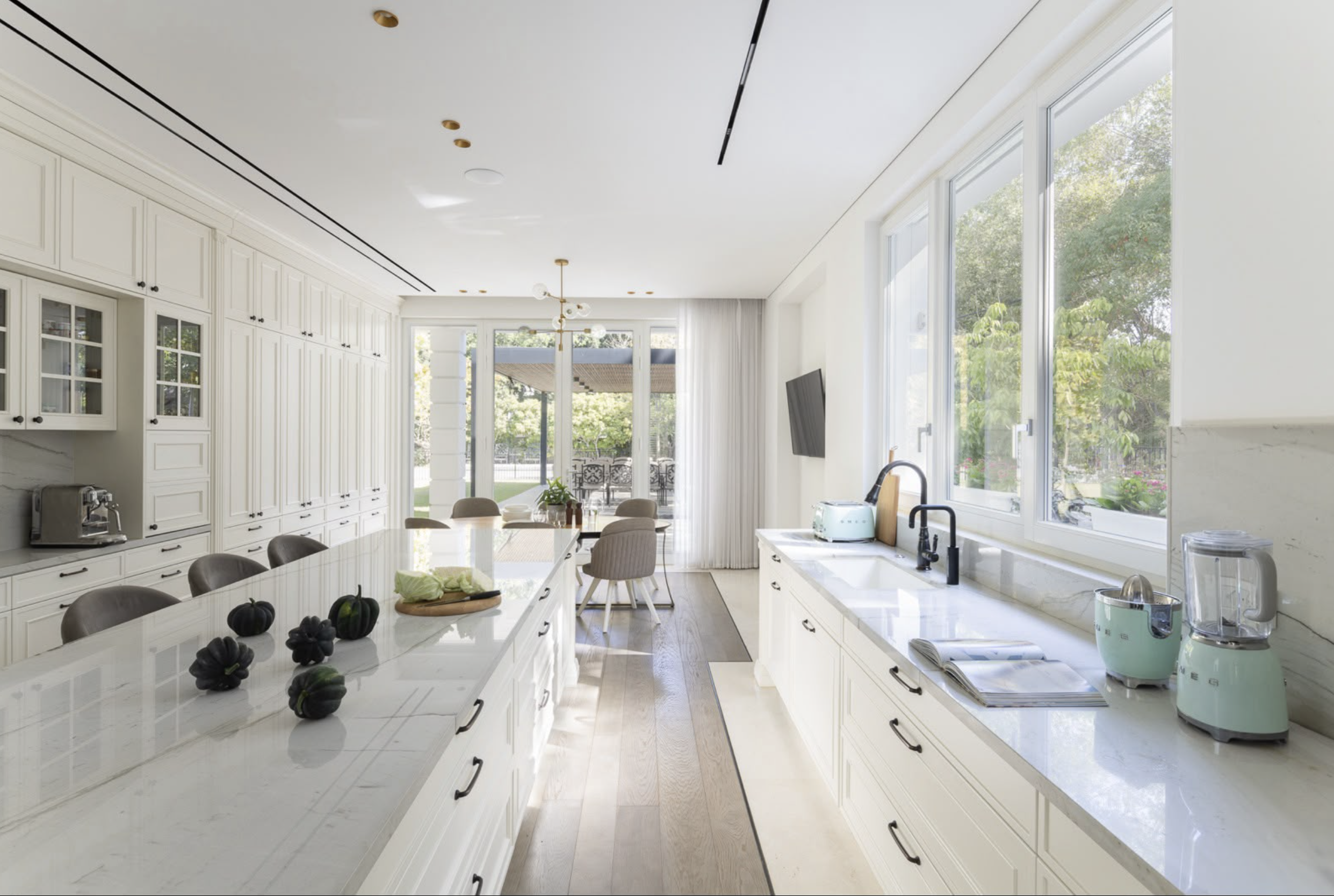 A bright, modern kitchen with white cabinets and countertops, large windows with a view of trees outside, a dining table with chairs, black pumpkins as decor, and various small appliances on the counters.