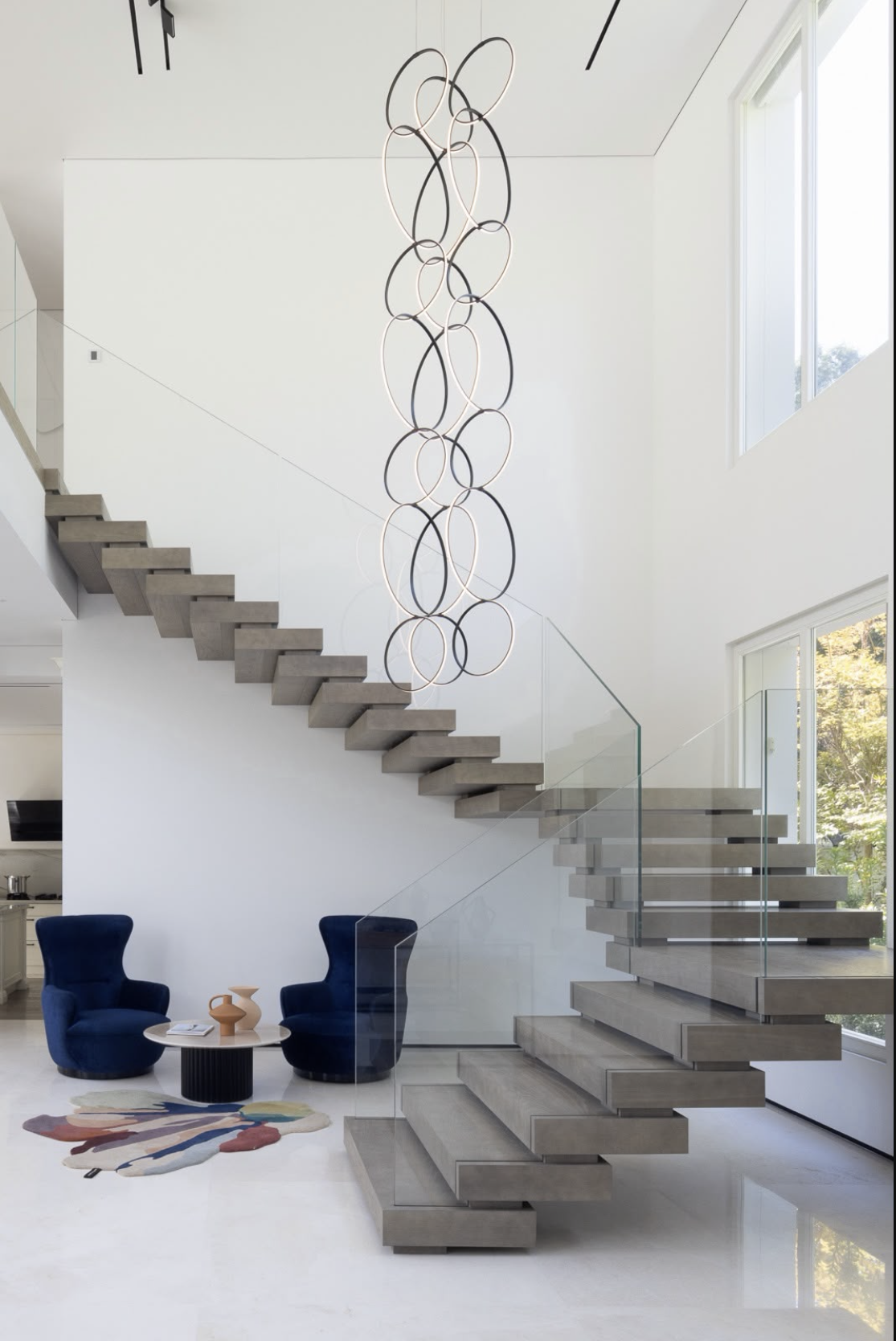 Modern interior staircase with floating wooden steps, glass railing, and a contemporary circular pendant light fixture hanging from the ceiling. The space includes a sitting area with two dark blue chairs, a small round black table with decorative va