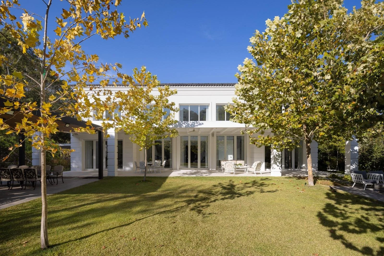A modern two-story white house with large windows, a spacious backyard with a well-maintained green lawn, several trees, and outdoor furniture including chairs and tables under a pergola and in the shade.