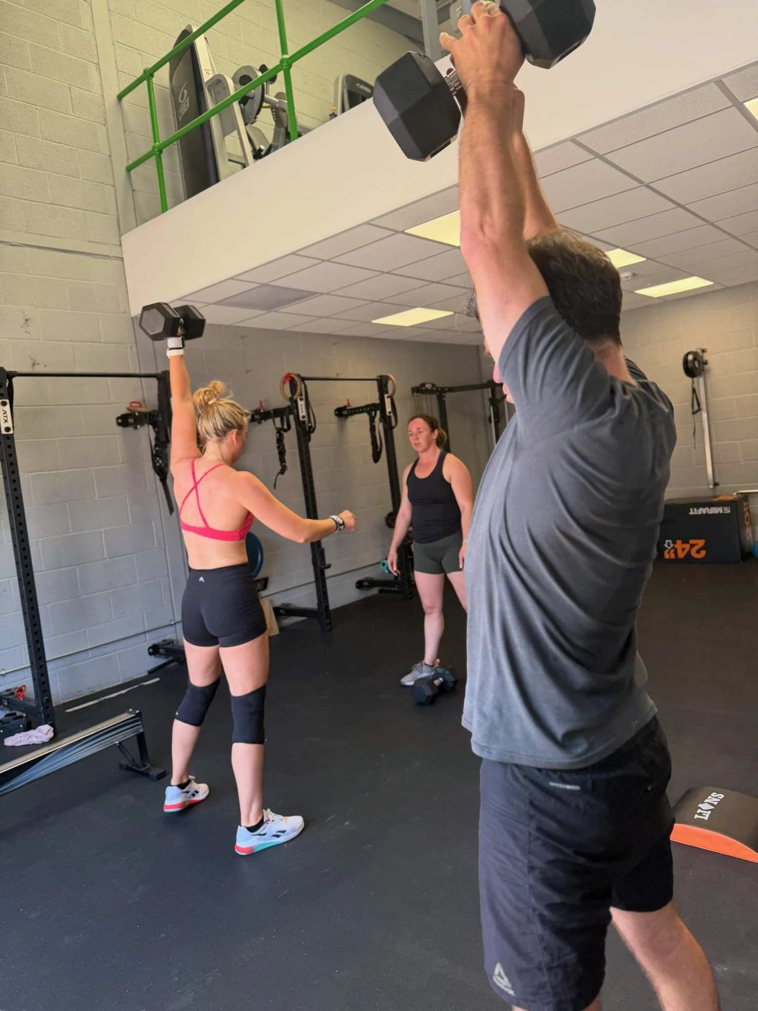 Three people working out inside a gym. One woman is lifting a dumbbell overhead, a man is performing a similar exercise, and another woman is standing nearby watching or coaching.