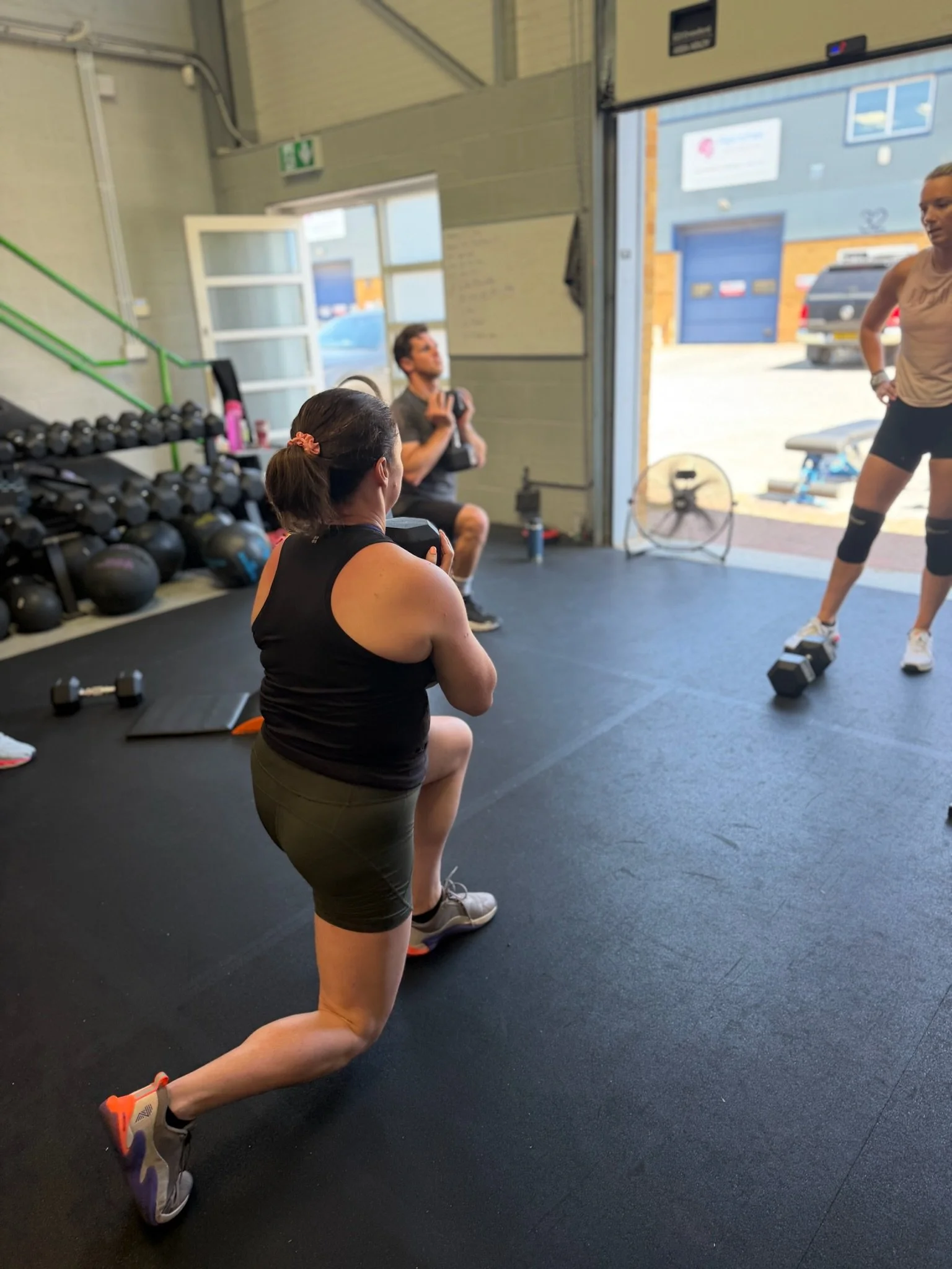 A woman in black athletic wear and sneakers performs a lunge exercise inside a gym, with other gym equipment and a partially open garage door in the background.