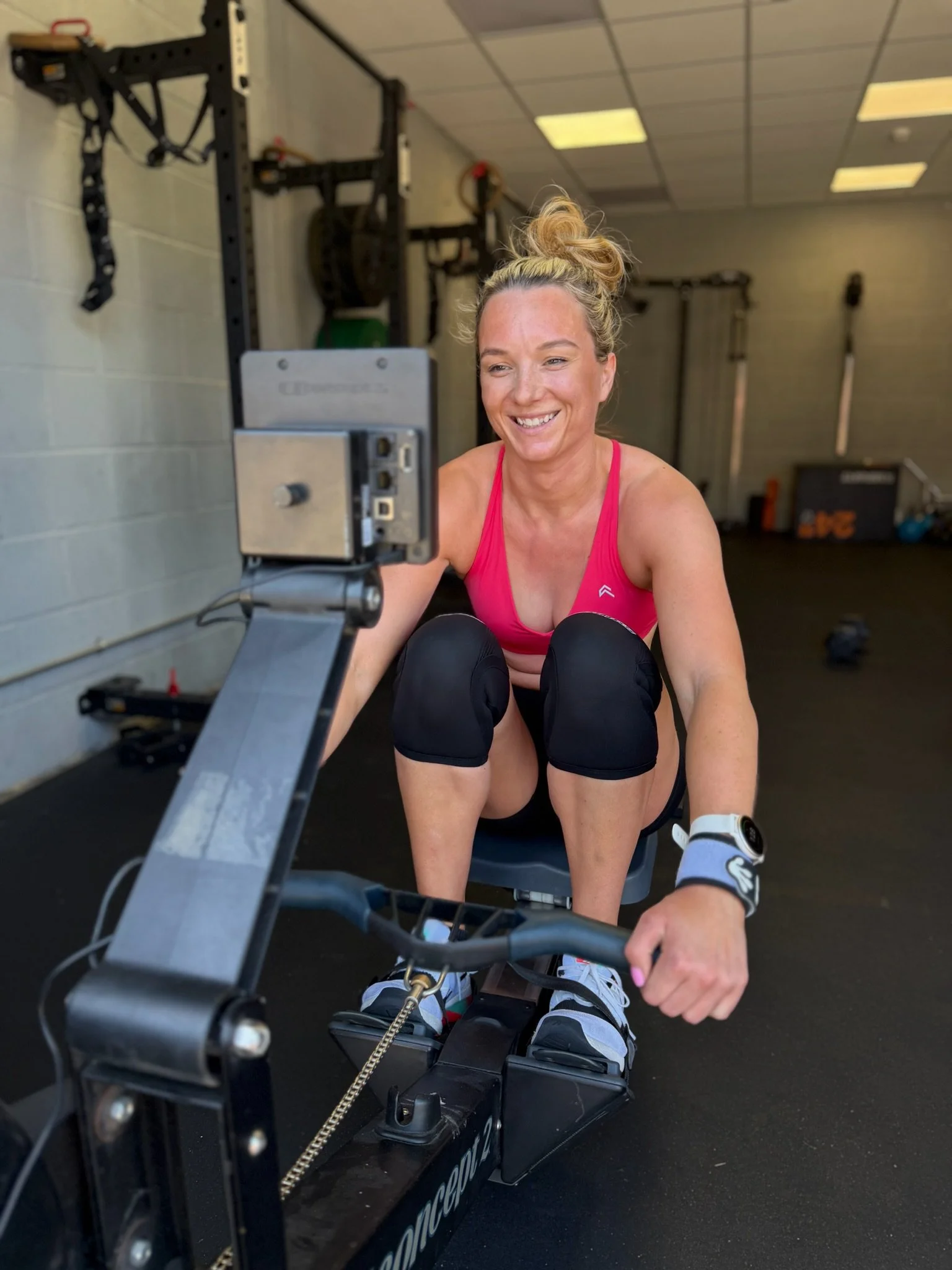 A woman with blonde hair tied in a bun is smiling while using a rowing machine in a gym. She has on a pink sports top, black athletic shorts, and is wearing a watch and knee straps. The gym has a black floor, a high ceiling with fluorescent lights, and equipment, including a wall-mounted squat rack with resistance bands and weights.