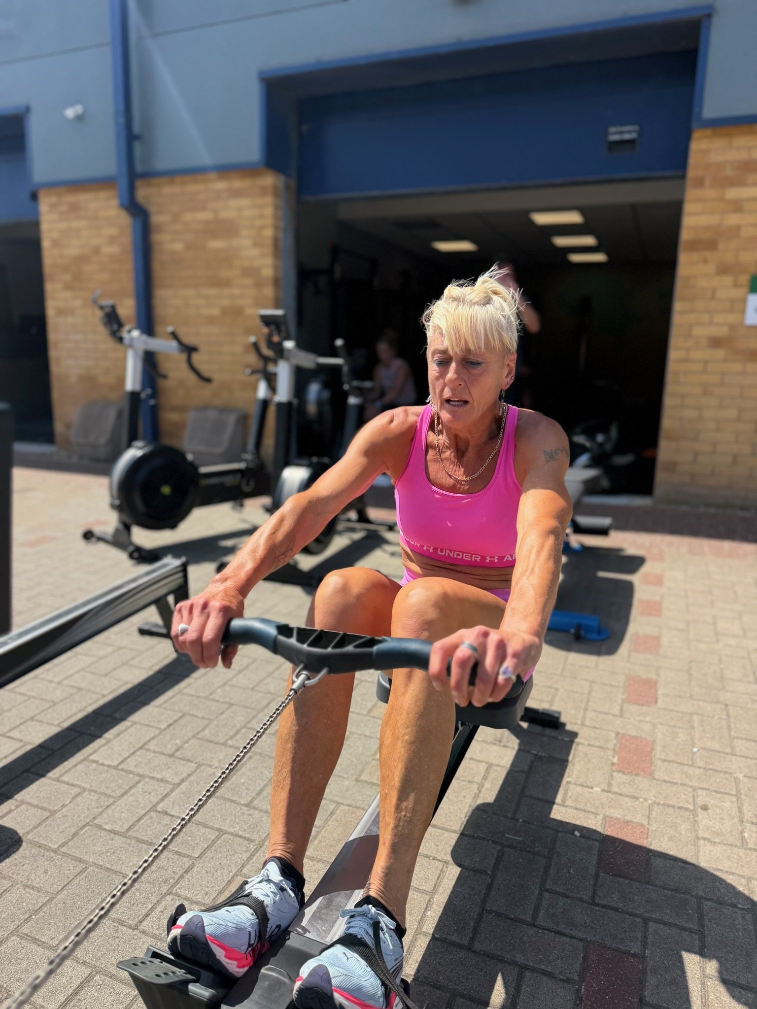 A woman wearing a pink athletic top and running shoes is using a rowing machine outdoors in front of a building with a brick wall and exercise equipment. She appears focused on her workout.