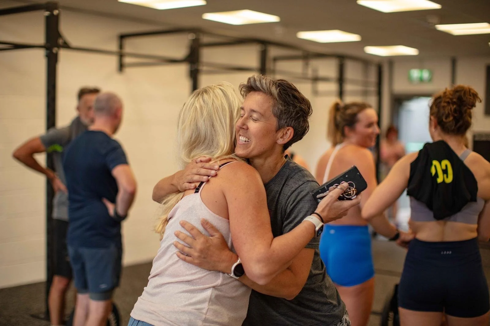 Group of diverse women and men at a gym, smiling and posing after a workout, with gym equipment and wall-mounted racks in the background.