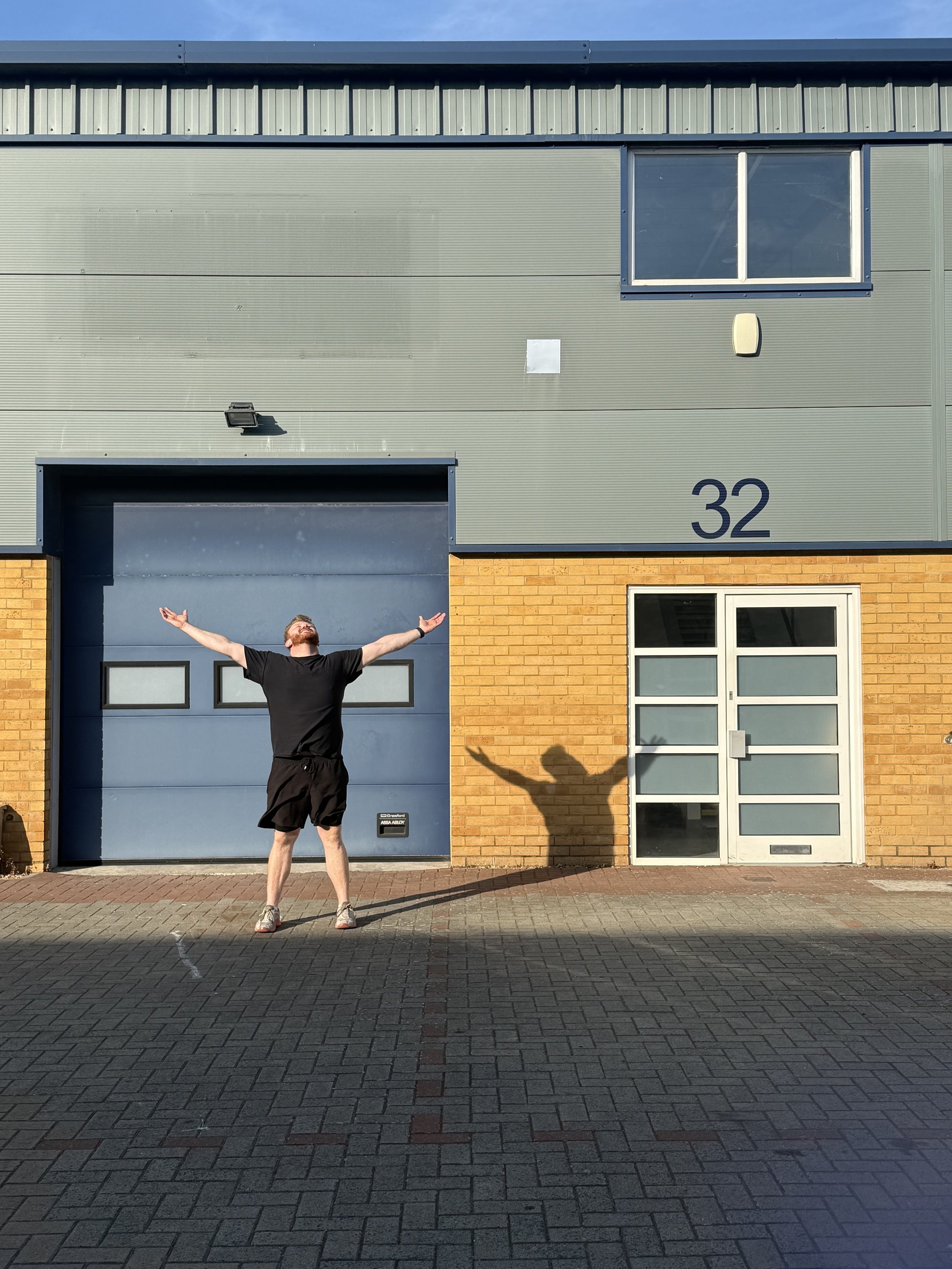 A man with a beard and short hair wearing a black T-shirt, black shorts, and sneakers standing outside in front of a building with a yellow brick lower section and a green upper section, arms outstretched, looking upwards, casting a shadow on the ground.