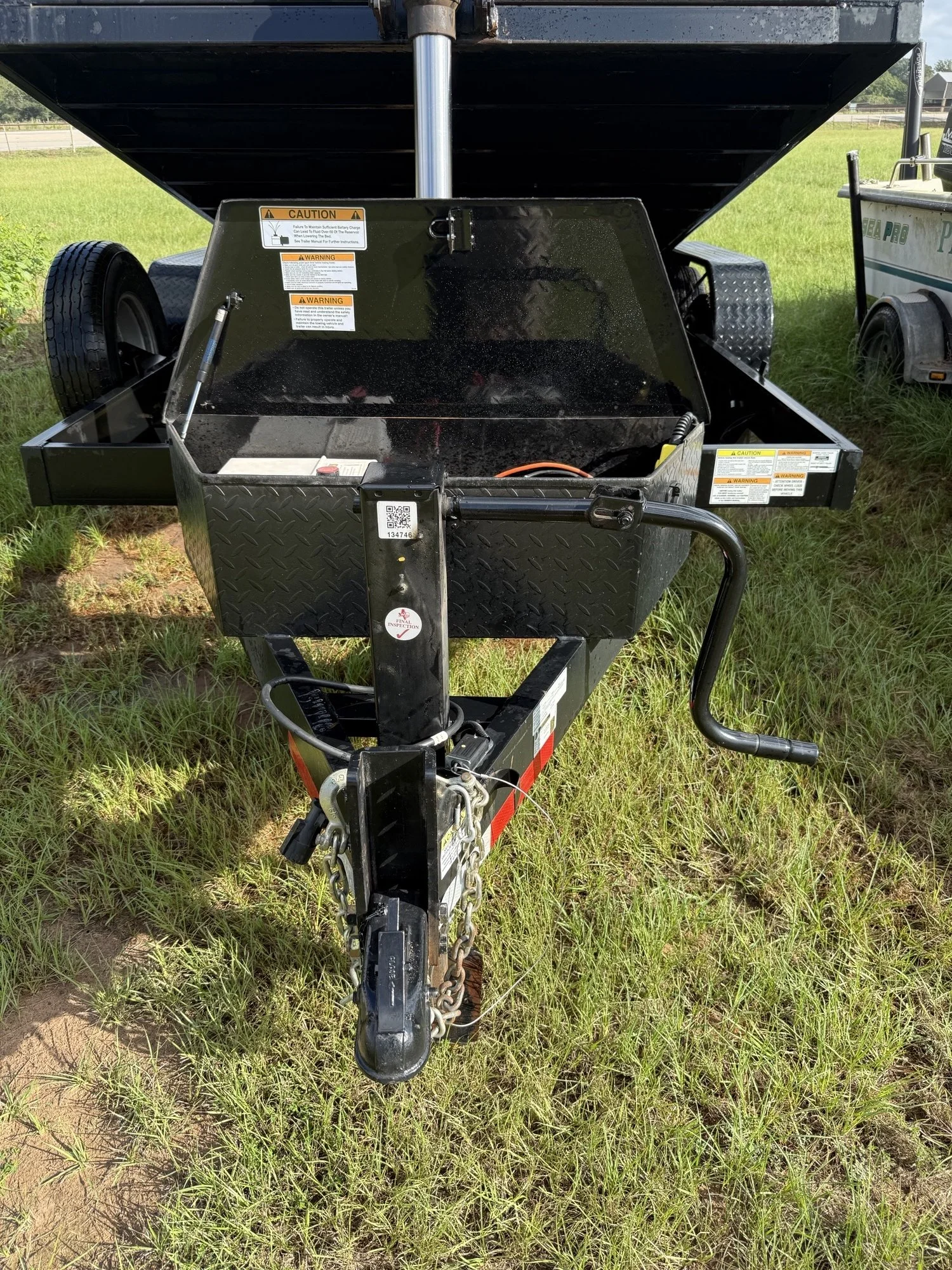 A trailer with a black metal cargo box on a grassy field, with a hitch and chains in the foreground, and some stickers and warning labels visible on the trailer.