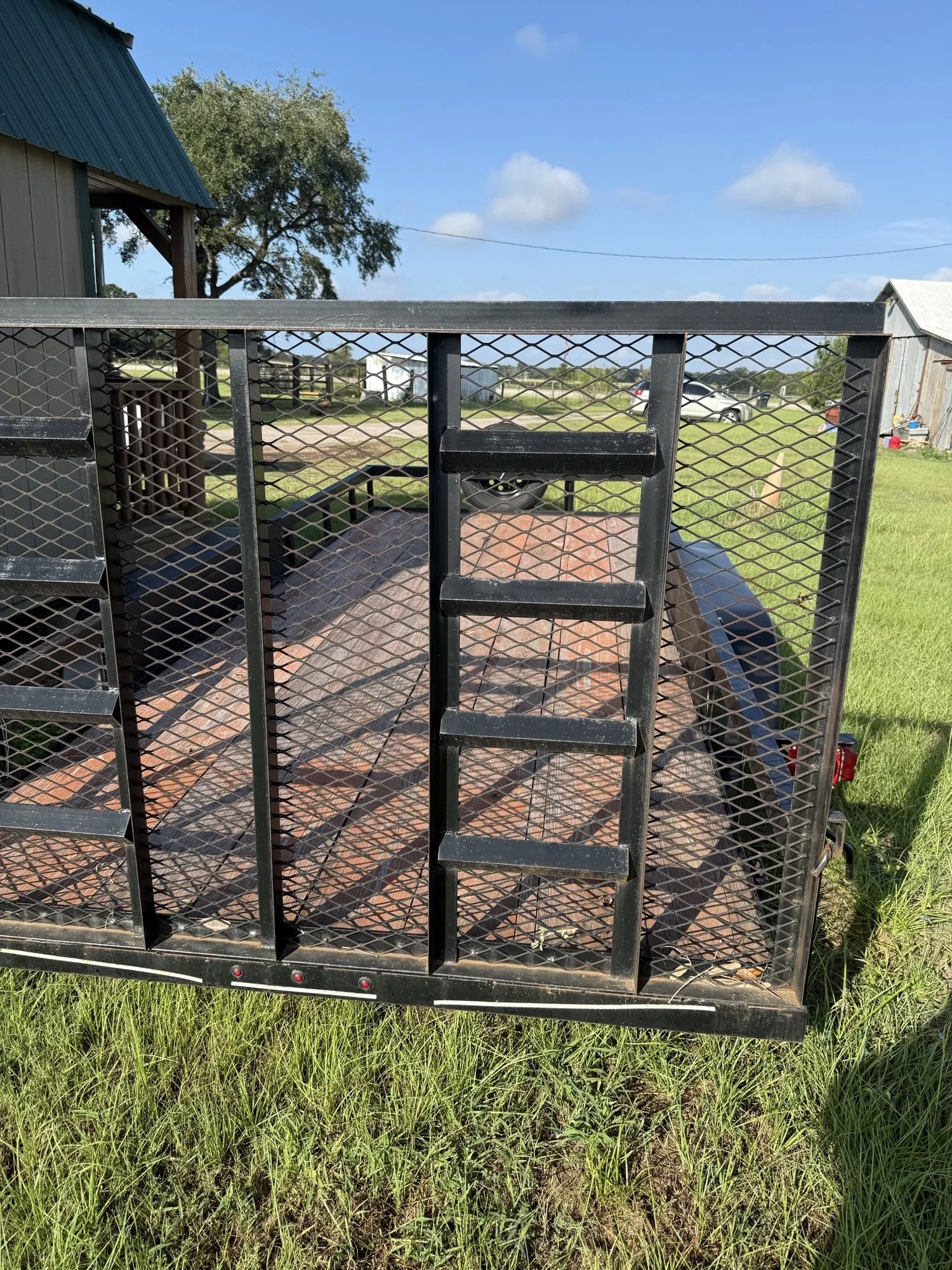 A black metal trailer with a grated side gate, parked on grassy land under a clear blue sky with scattered clouds. A barn and a few cars are visible in the background.