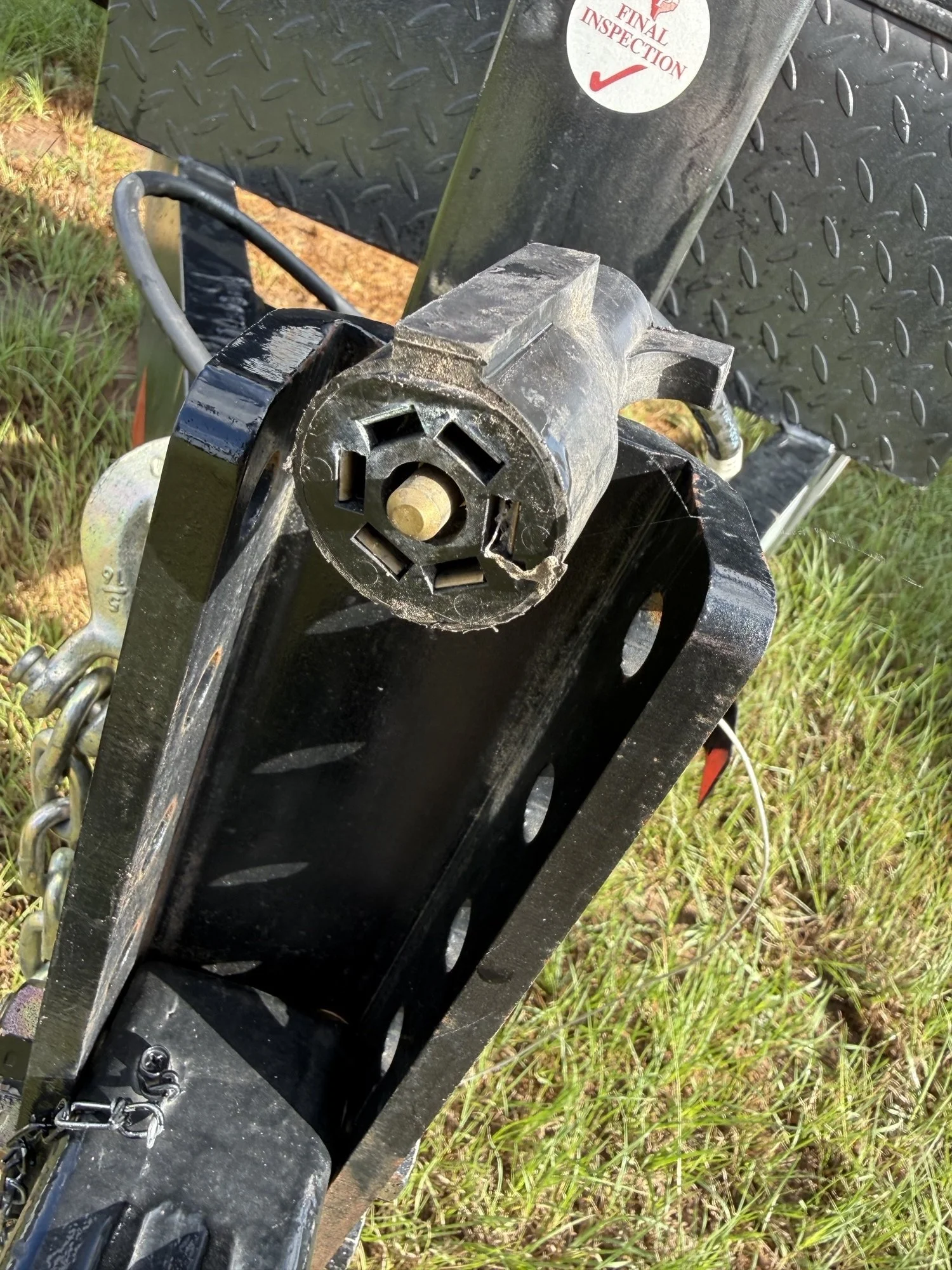 Close-up of a trailer hitch and safety chains attached to a black trailer with a metal diamond plate side panel, outdoors on grass and dirt.