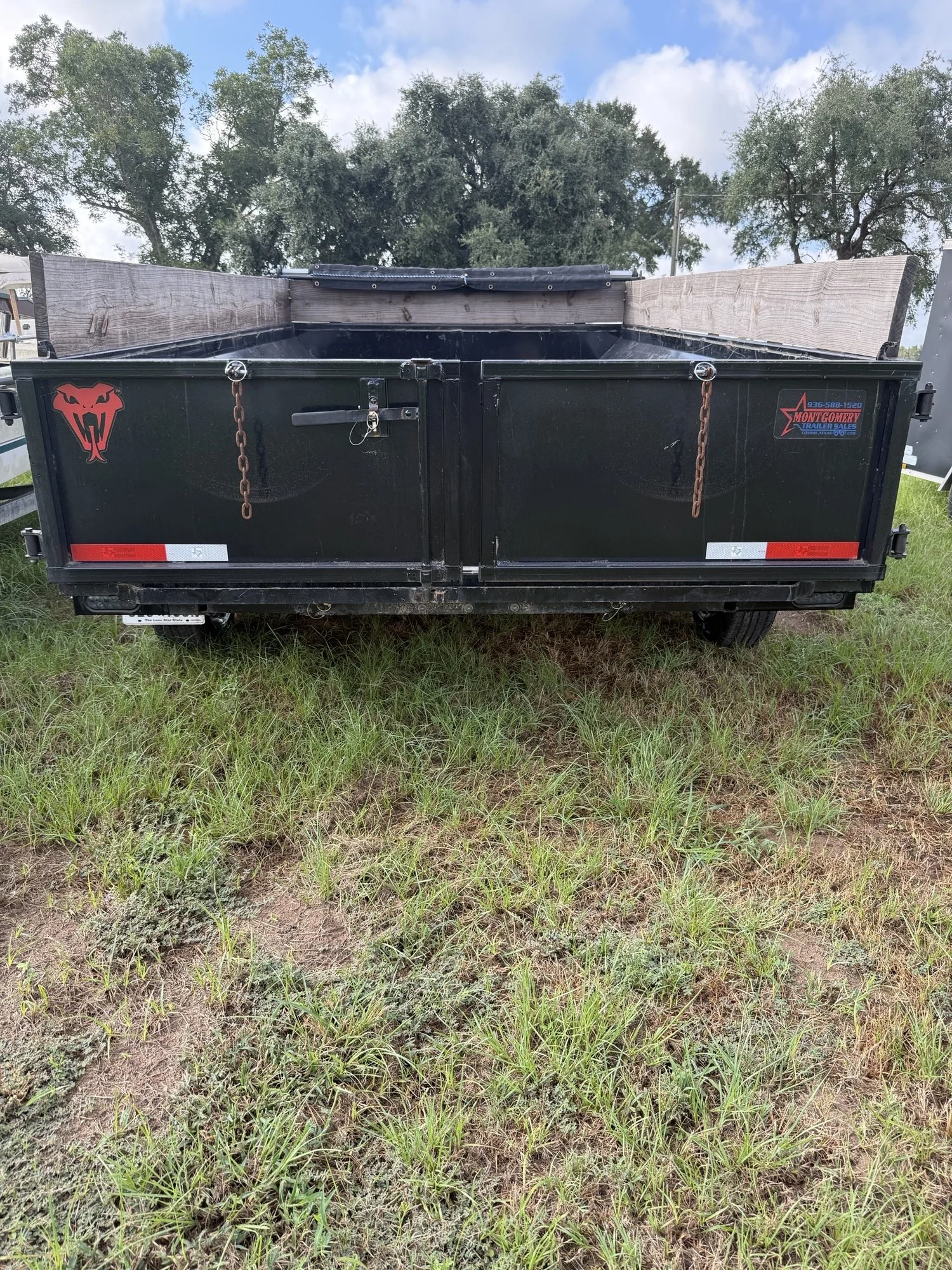 A black utility trailer with wooden side panels, parked on grass under a partly cloudy sky, with trees visible in the background.