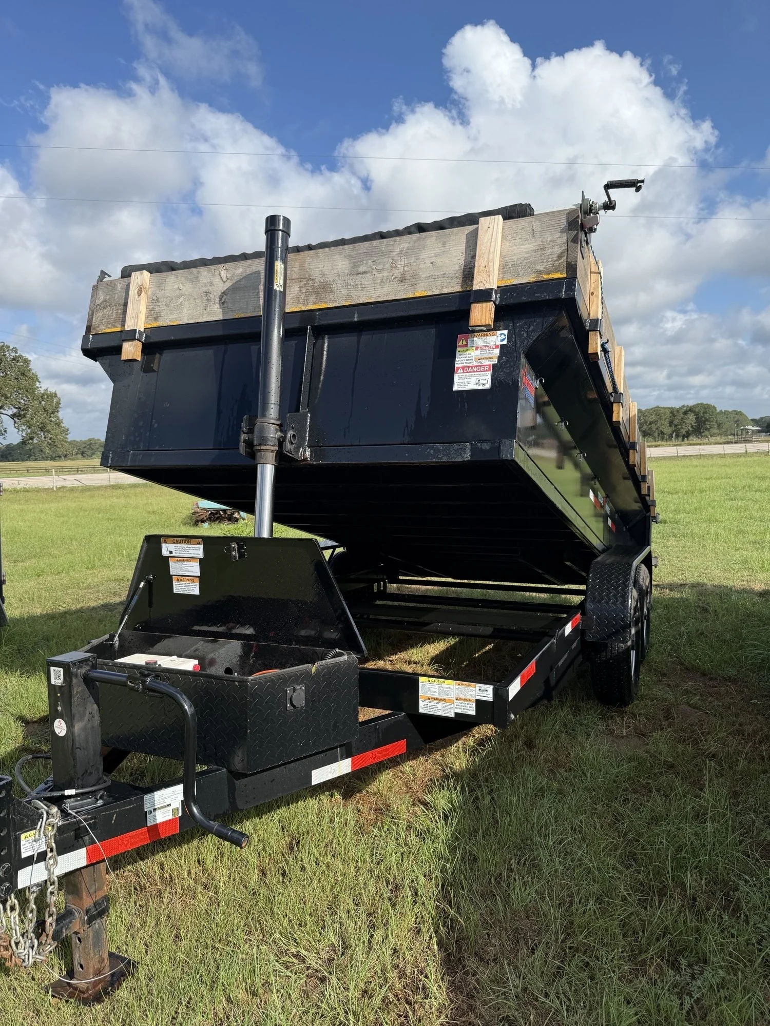 A black flatbed trailer with a hydraulic lift, parked on grass, under a partly cloudy sky.