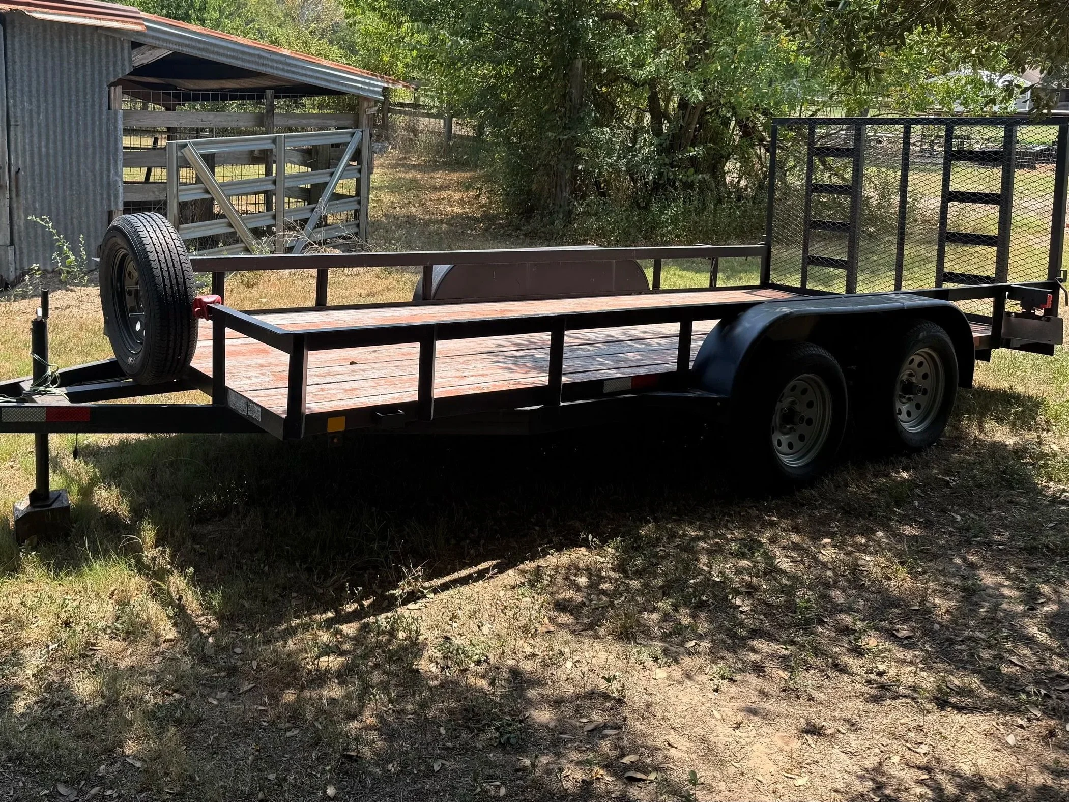 A flatbed trailer with a wooden deck parked on a grassy area next to a shed and trees, with a metal gate in the background.