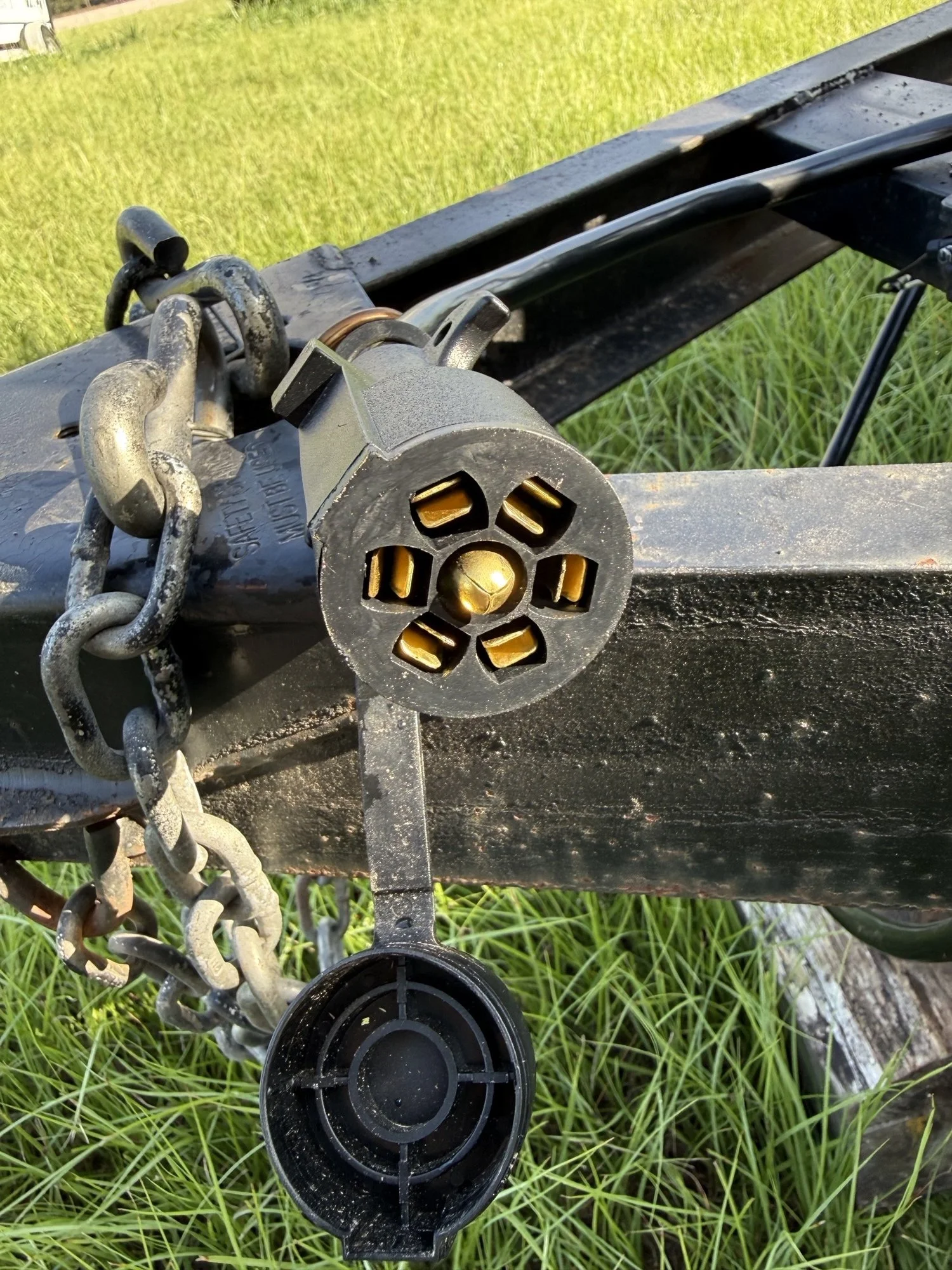 Close-up of a black trailer hitch with a locking mechanism, chains, and a socket, set against green grass.