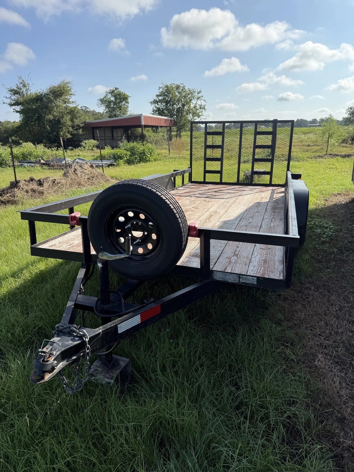 An empty flatbed trailer with a spare tire mounted on the front, parked on green grass in a rural area with trees and a small building in the background.
