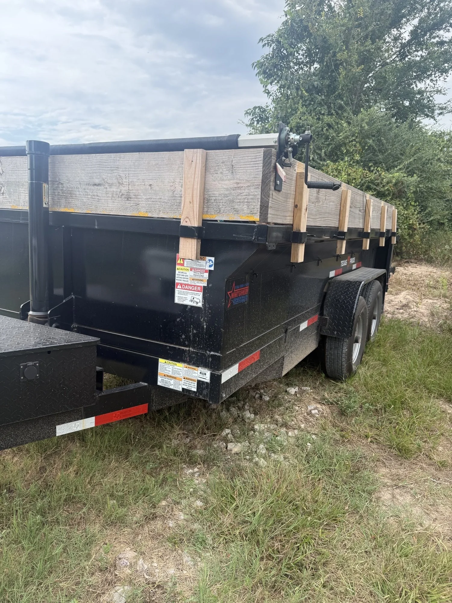 A black flatbed trailer with a wooden fence on top, parked on grass with trees in the background, a cloudy sky overhead.