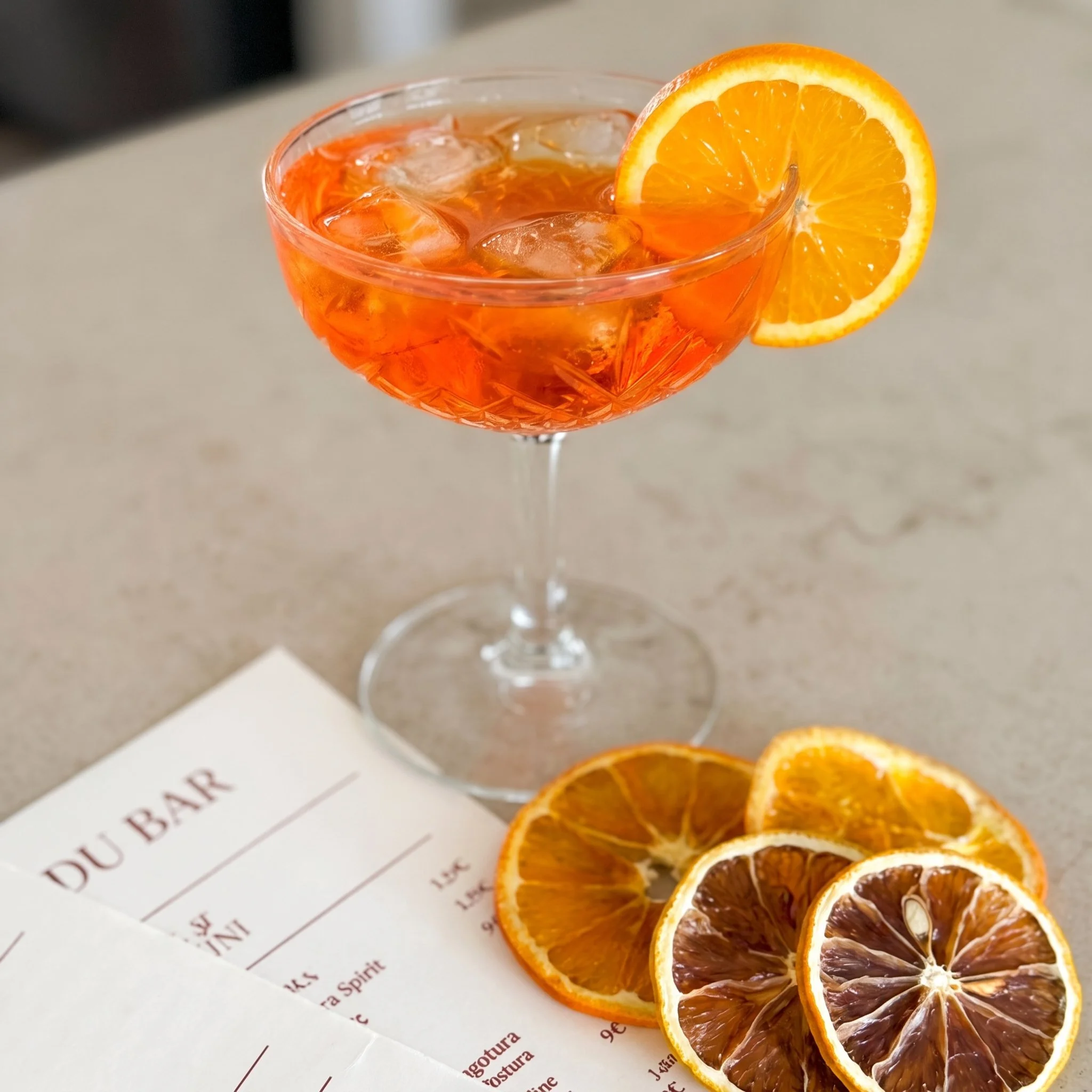 Un verre à cocktail rempli de boisson orange avec des glaçons, décoré d'une tranche d'orange sur le rebord, posé sur une surface claire. À l'arrière-plan, une feuille de menu est partiellement visible.