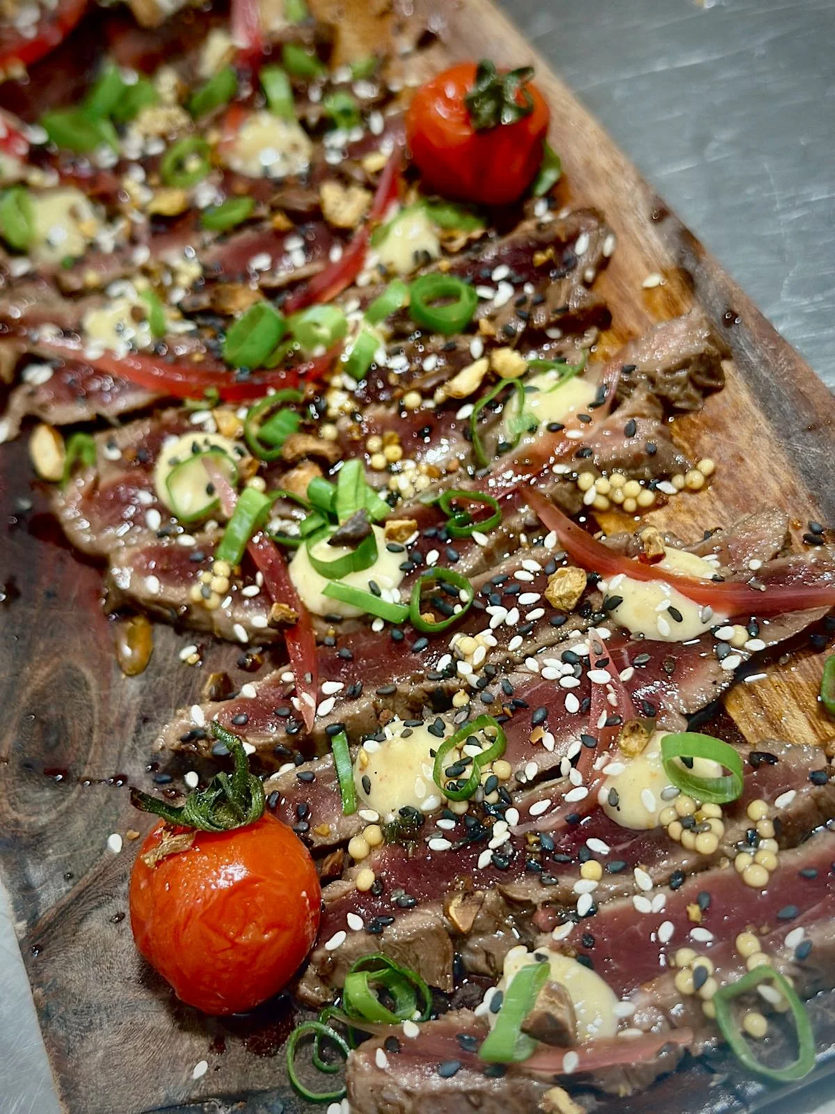 Une planche en bois avec une tartare de viande garnie de tomates cerises, oignons verts, graines de sésame, et diverses épices.