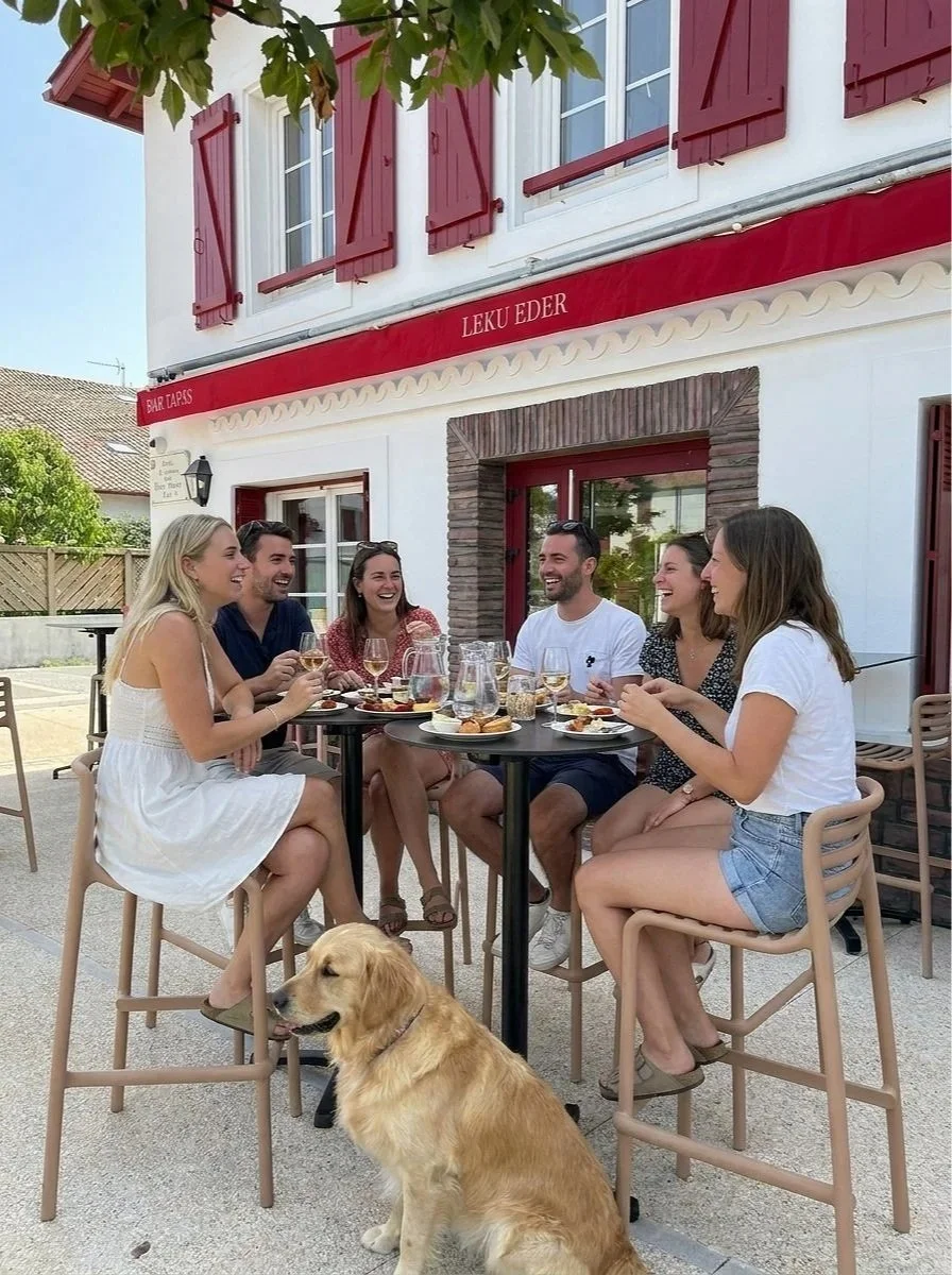 Groupe de six personnes assises autour d'une table en terrasse, partageant un repas et riant. Un chien golden retriever est assis à côté d'eux. Contexte d'une façade de bâtiment avec volets rouges, sous un ciel ensoleillé.