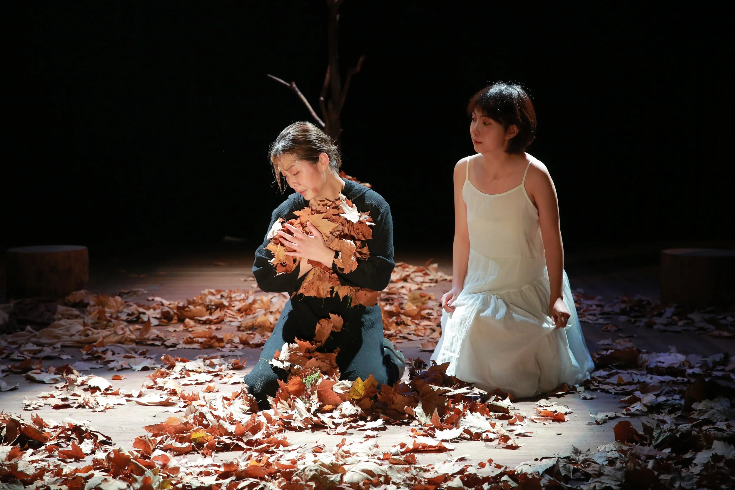 Two women are on a stage surrounded by fallen autumn leaves. One woman, kneeling with eyes closed, is holding a bouquet of leaves. The other woman, in a white dress, is kneeling beside her, looking at her with a gentle expression. The background is dark with a tree branch visible, suggesting an outdoor or forest setting.