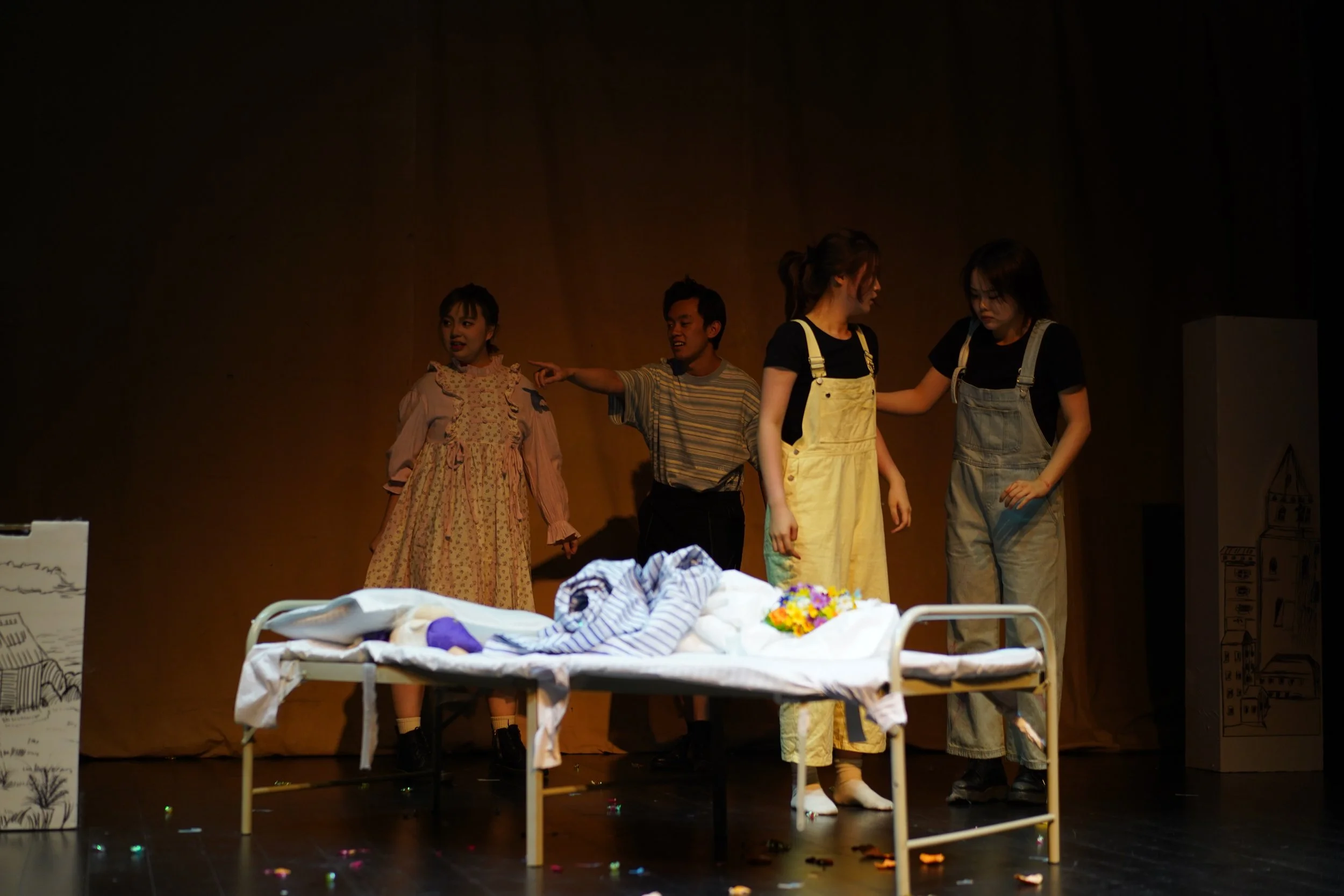 Four young actors on stage during a theatrical performance, with a hospital bed, drawings, and cardboard boxes as props.