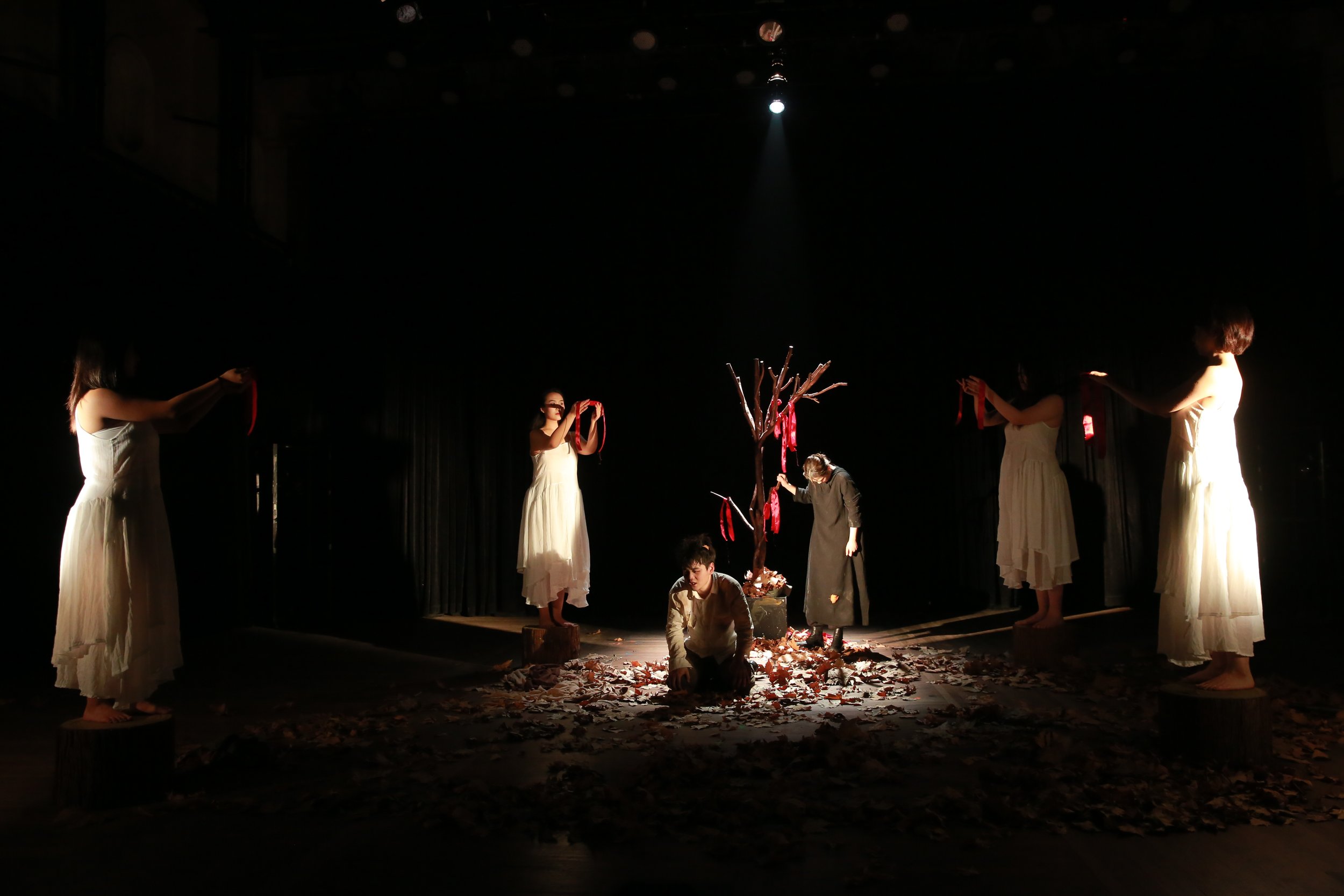 The image depicts a theatrical stage with several actors dressed in white and dark clothing, standing around a central tree-like prop. The stage is dimly lit, with a spotlight focusing on the performers and the ground covered with leaves, creating a dramatic atmosphere.