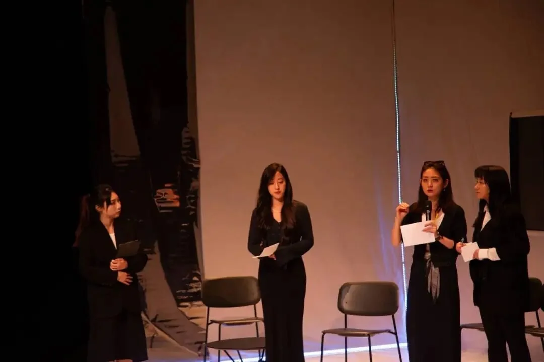 Four women dressed in black professional attire, standing on a stage with three chairs, engaged in a discussion or presentation.