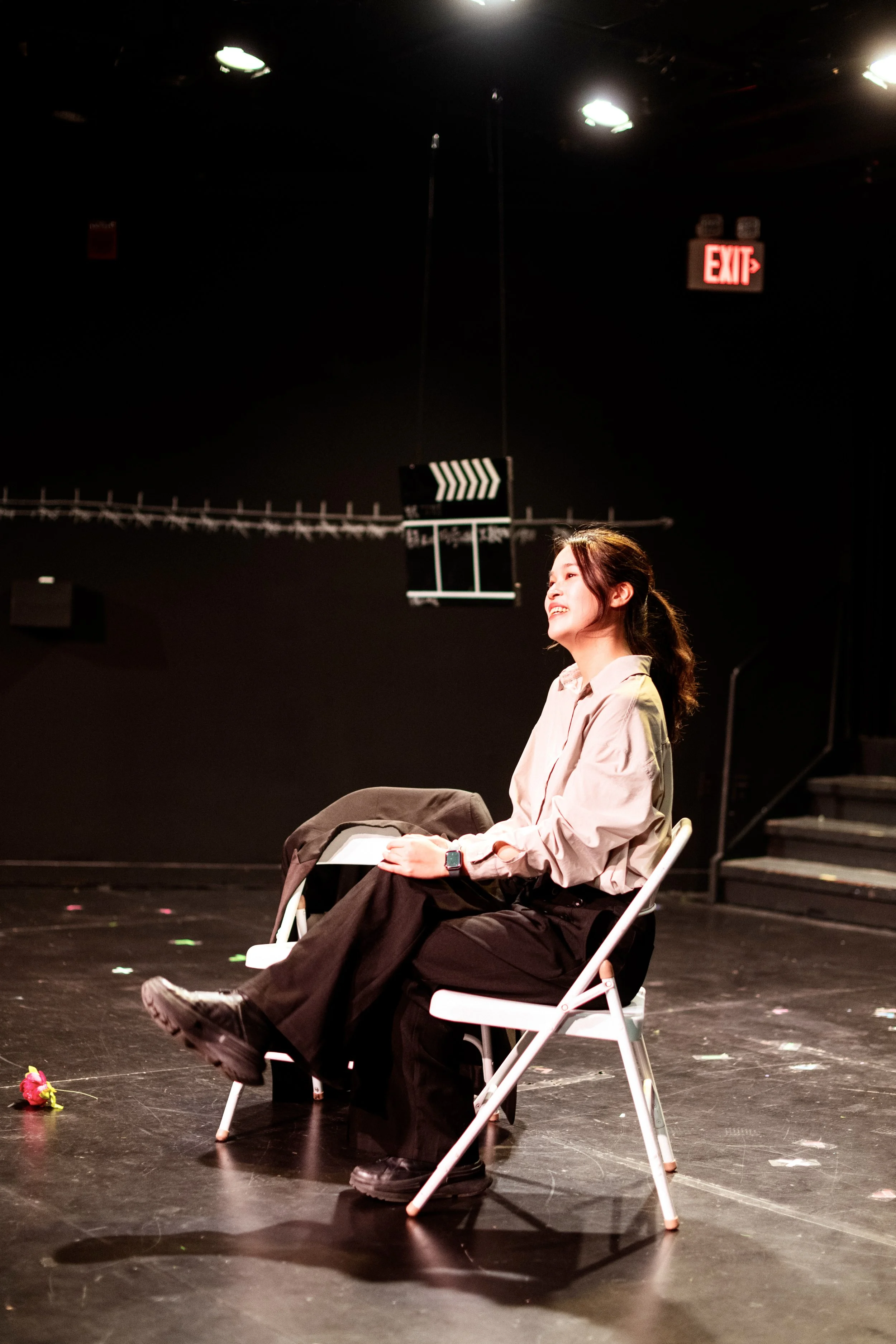 A young woman sitting on a white folding chair on a stage, smiling, with a black jacket on her lap, a smartwatch on her left wrist, and a flower on the floor in front of her. The stage background is dark with an exit sign and stage lighting.