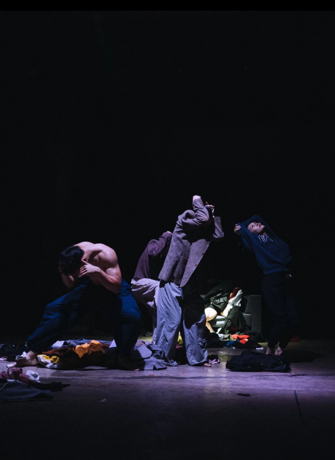 Three people are in a dark room with scattered clothes on the floor. One person is shirtless, crouching, and covering their face. The second person, in the center, is standing with their head tilted back, wearing a gray jacket, gray pants, and a purple head covering. The third person, on the right, is standing with their head tilted back and hands behind their head, dressed in a dark hoodie and pants.
