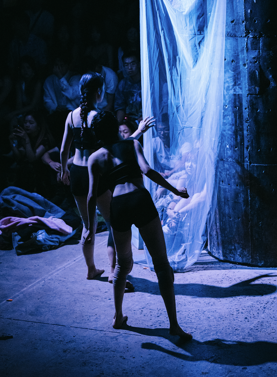 Two women in black dance costumes perform in front of a sheer white curtain with an audience seated in darkness in the background