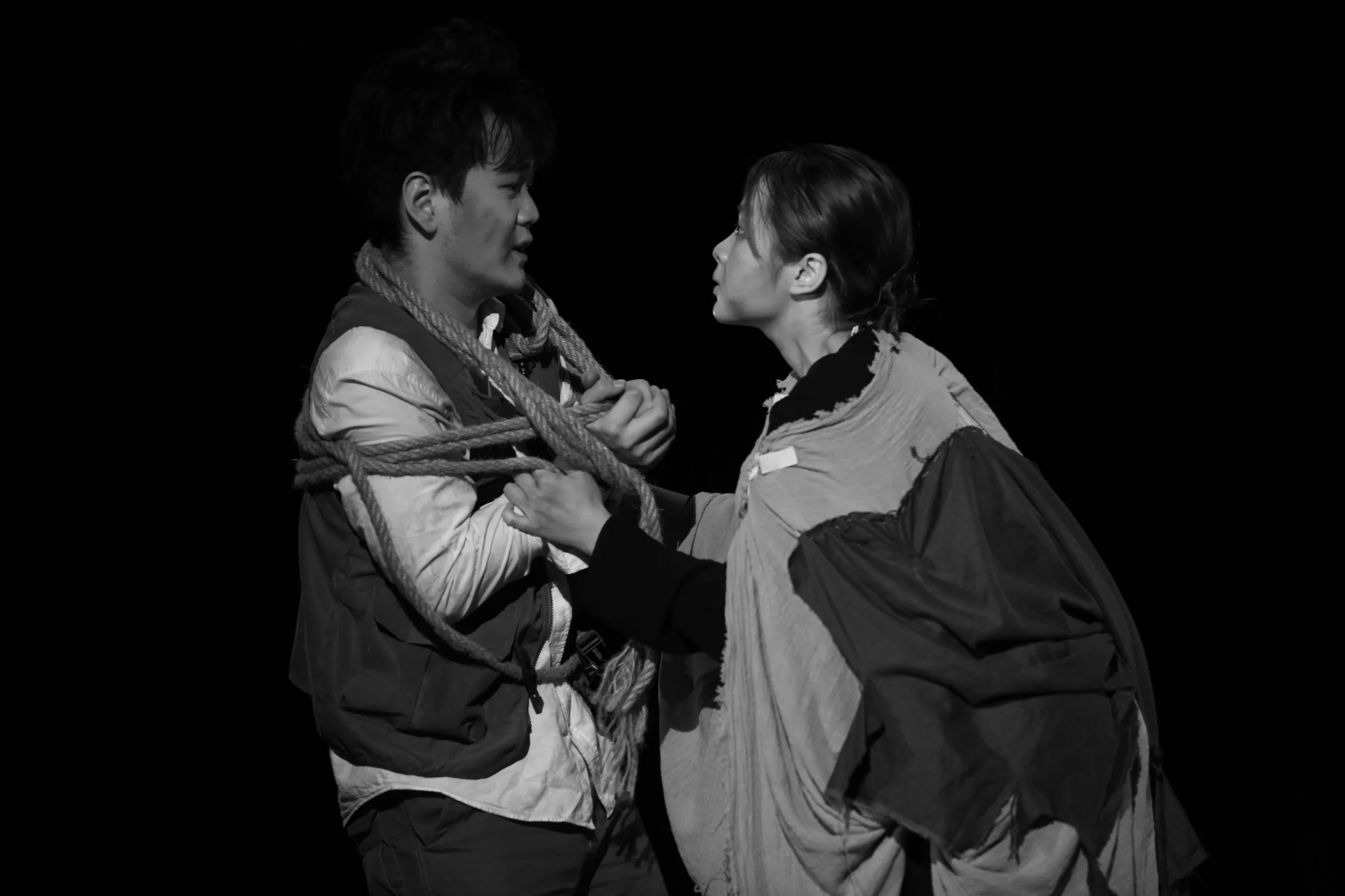 Two people facing each other with intense expressions, holding hands, while one person is wrapped in ropes, against a black background.