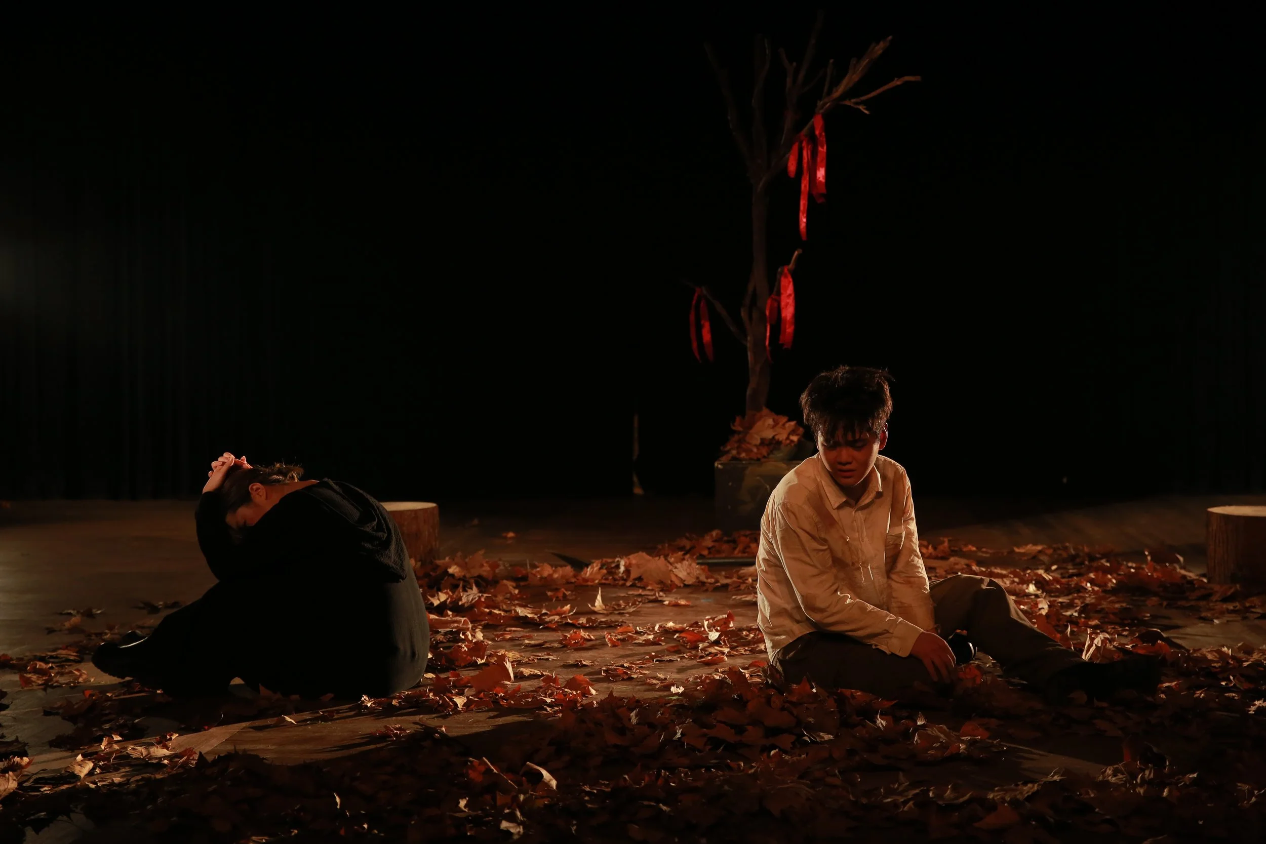 Two young individuals sitting on a stage covered with fallen leaves, with a small tree decorated with red ribbons in the background, under dim stage lighting.