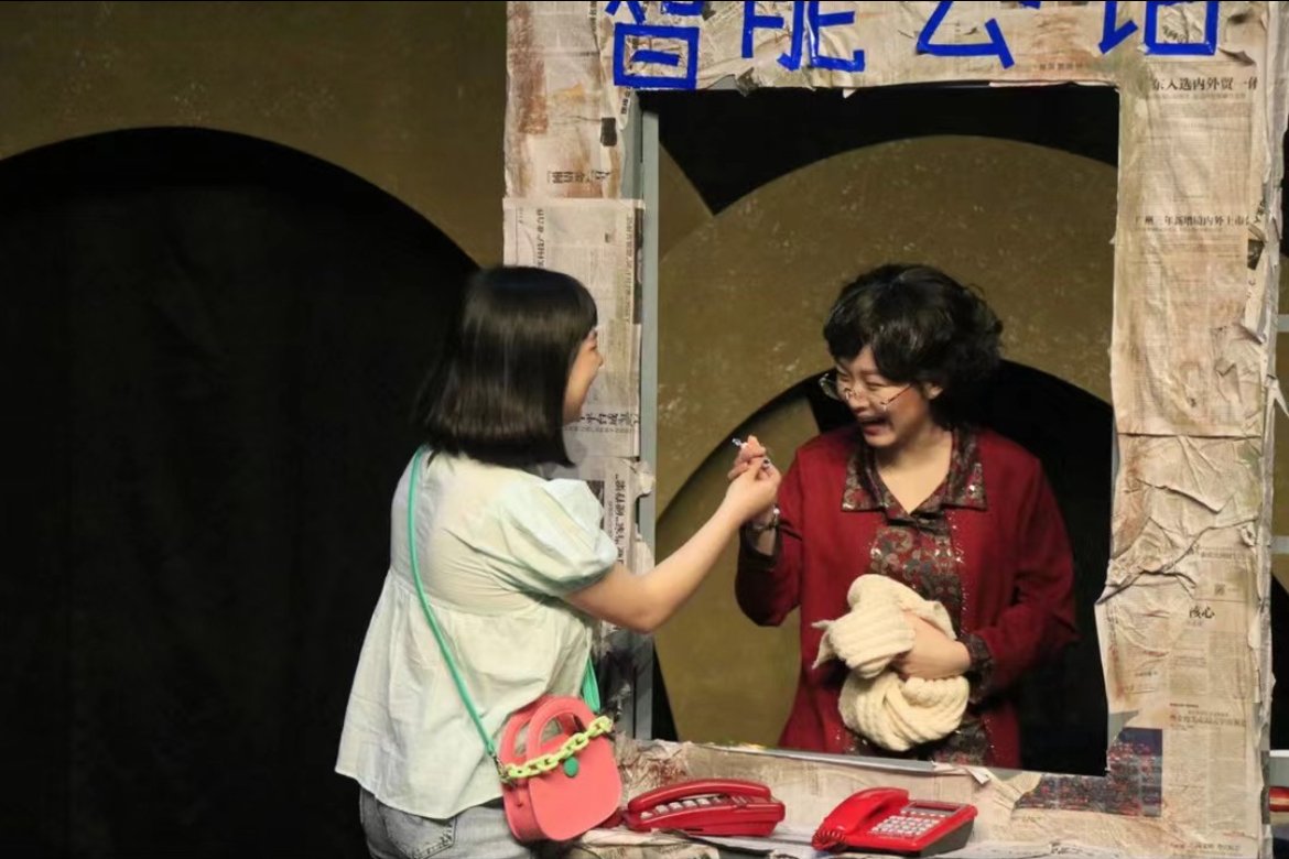 A young girl with a pink purse and a woman in a red jacket exchanging a smile through a window booth.