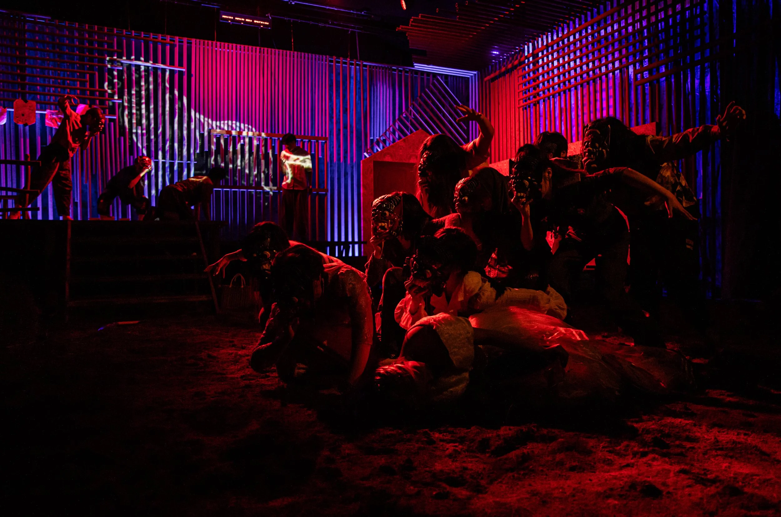 The image shows a theatrical scene with multiple performers dressed in masks and costumes, in a dark setting illuminated by red and purple stage lighting, with a backdrop of wooden slats and stairs.