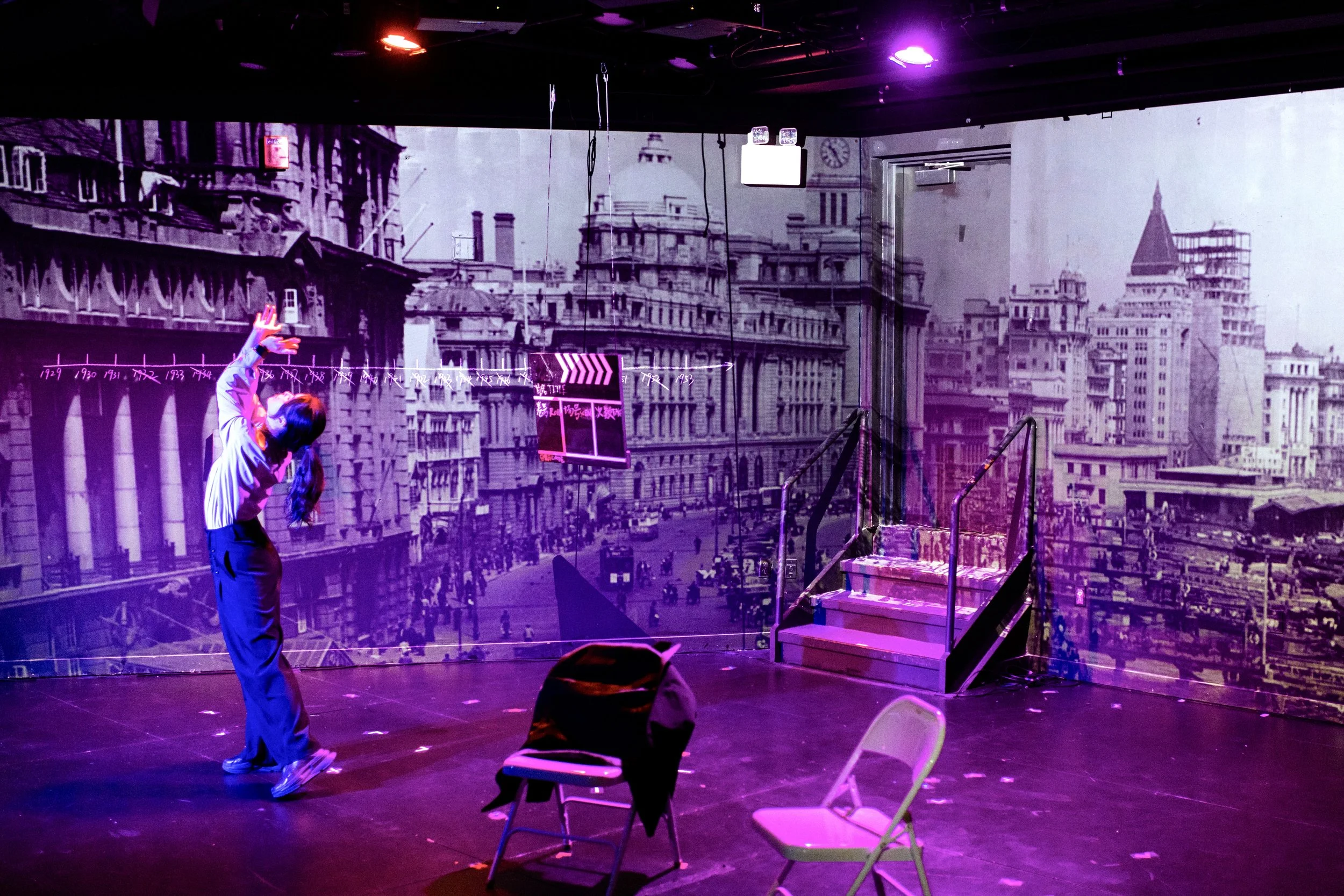 A stage set with a black and white cityscape mural in the background, purple stage lighting, a woman with dark hair in a ponytail pointing upwards, and several chairs on the stage.