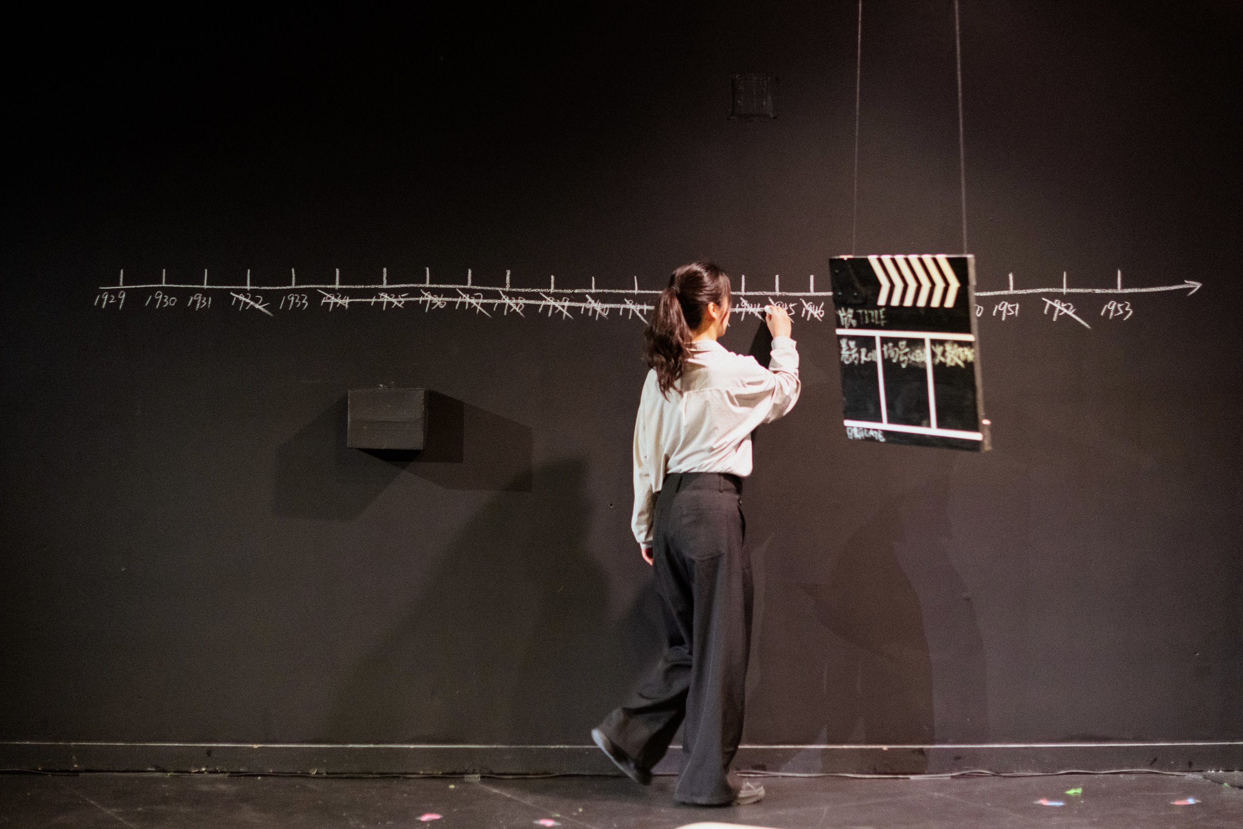 A woman with long dark hair tied back, wearing a light-colored long-sleeve shirt and black pants, stands on a stage writing on a large black chalkboard with a white marker. The chalkboard features a timeline with years labeled from 1929 through 1953. The stage background is black with some stage equipment and a mirror reflecting the woman.