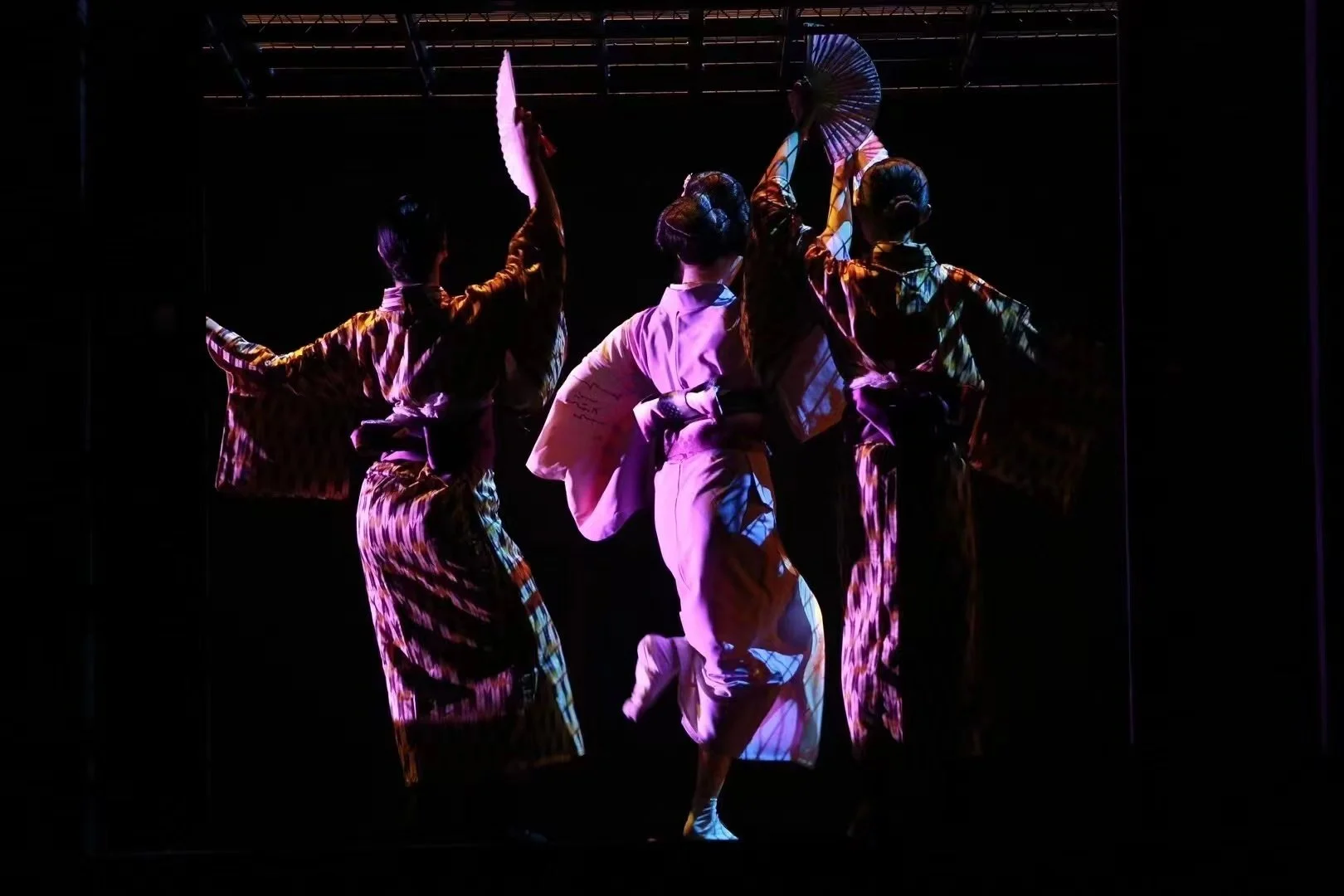 Four traditional Japanese dancers in colorful kimonos perform on stage, illuminated by purple and blue lighting, with fans and umbrellas as props.